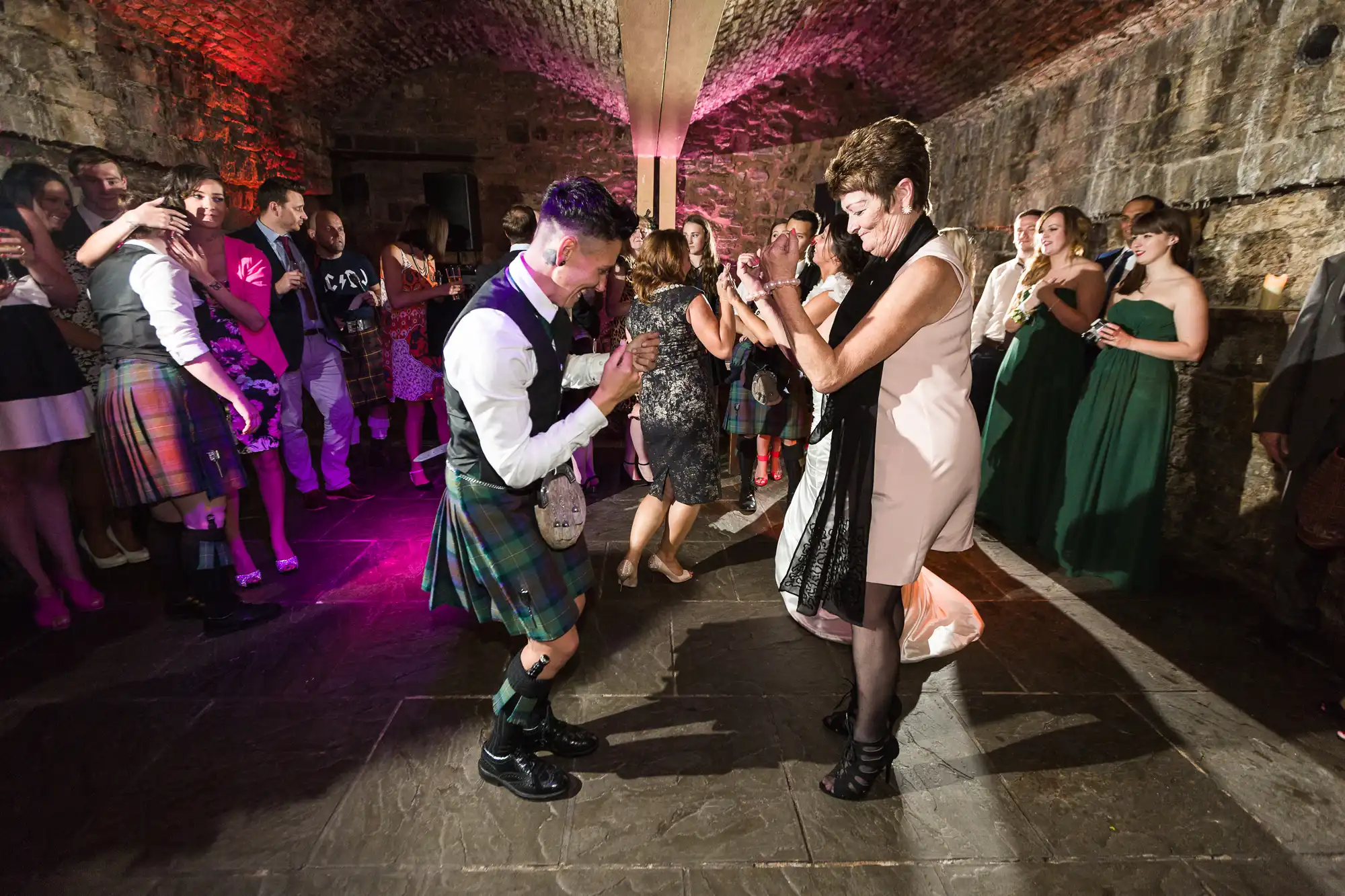 A lively group of people at a stone-walled venue dance together. In the foreground, a man in a kilt and a woman in a dress clap and smile, whilst other guests, some in formal attire, watch and enjoy the celebration under coloured lights.