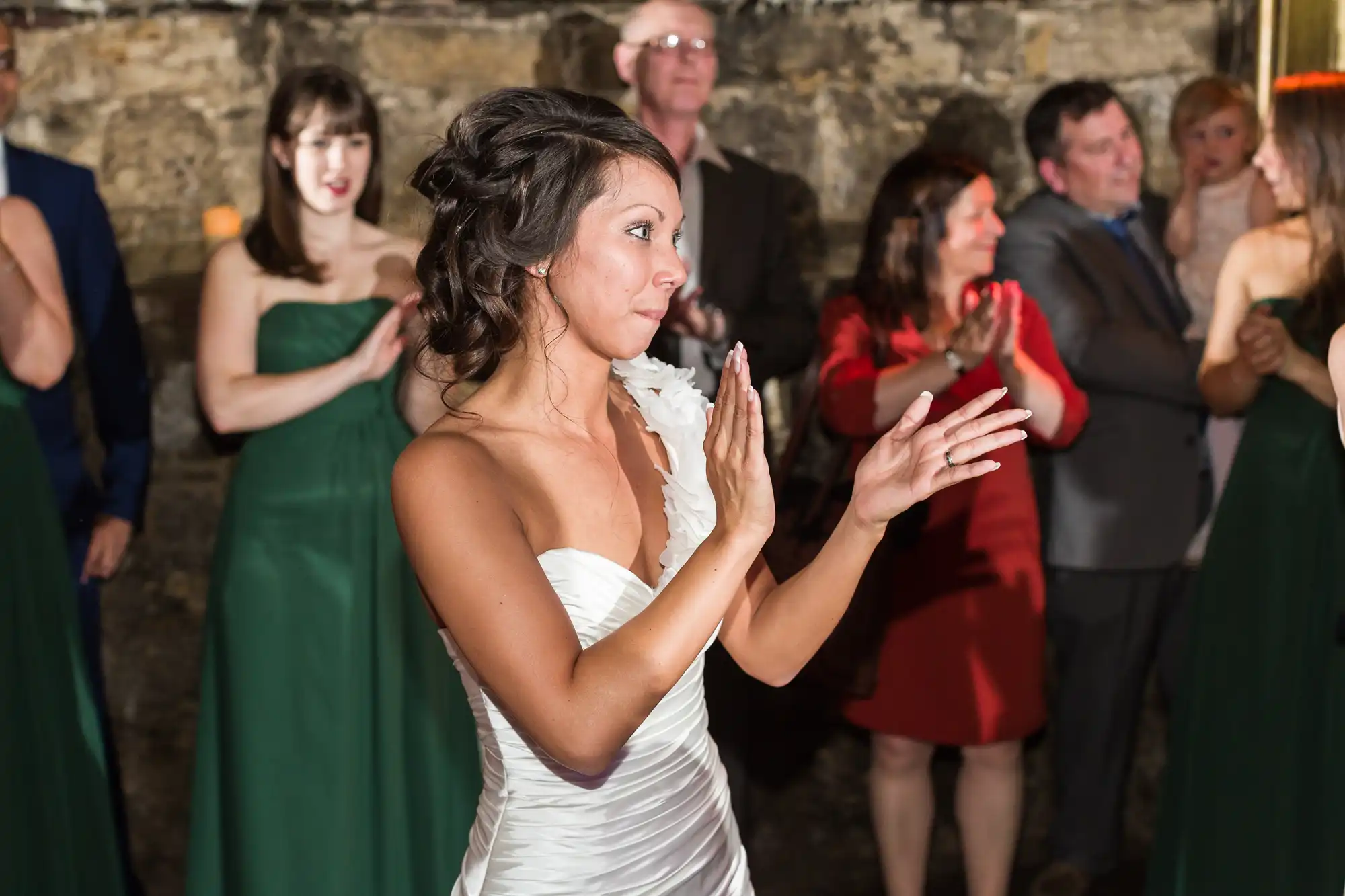 A bride in a white dress claps her hands, surrounded by guests. The guests, some in green dresses and one in a red dress, stand against a stone wall, smiling and clapping. The atmosphere is festive, likely at a wedding celebration.