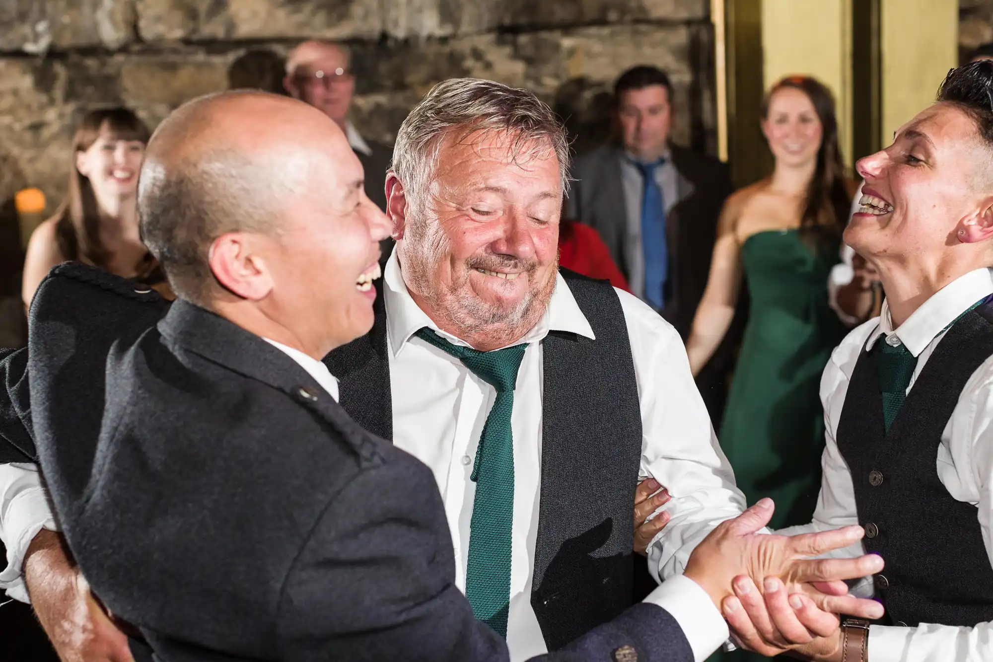 Three men in formal wear, including waistcoats and ties, are laughing and holding hands while dancing at an indoor event. The older man in the centre looks joyful. Other guests, some dressed up, are smiling and watching in the softly lit background.