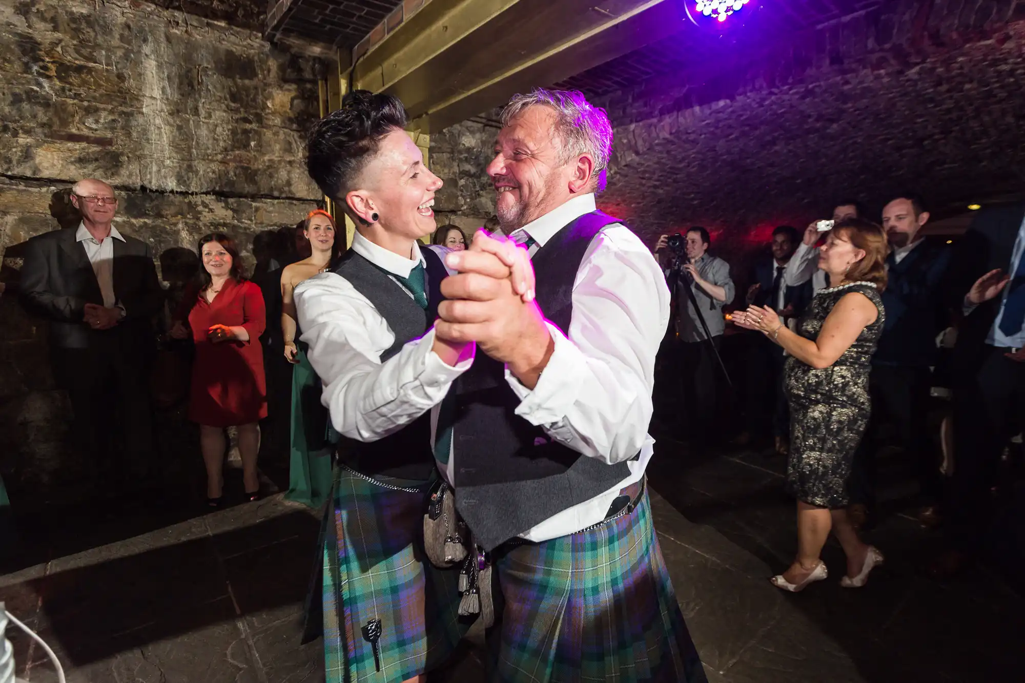 Two men in kilts joyfully dance together at an indoor celebration, smiling and making eye contact. Guests stand around them clapping and watching, some dressed formally. The venue has stone walls and beams, with purple party lights above.