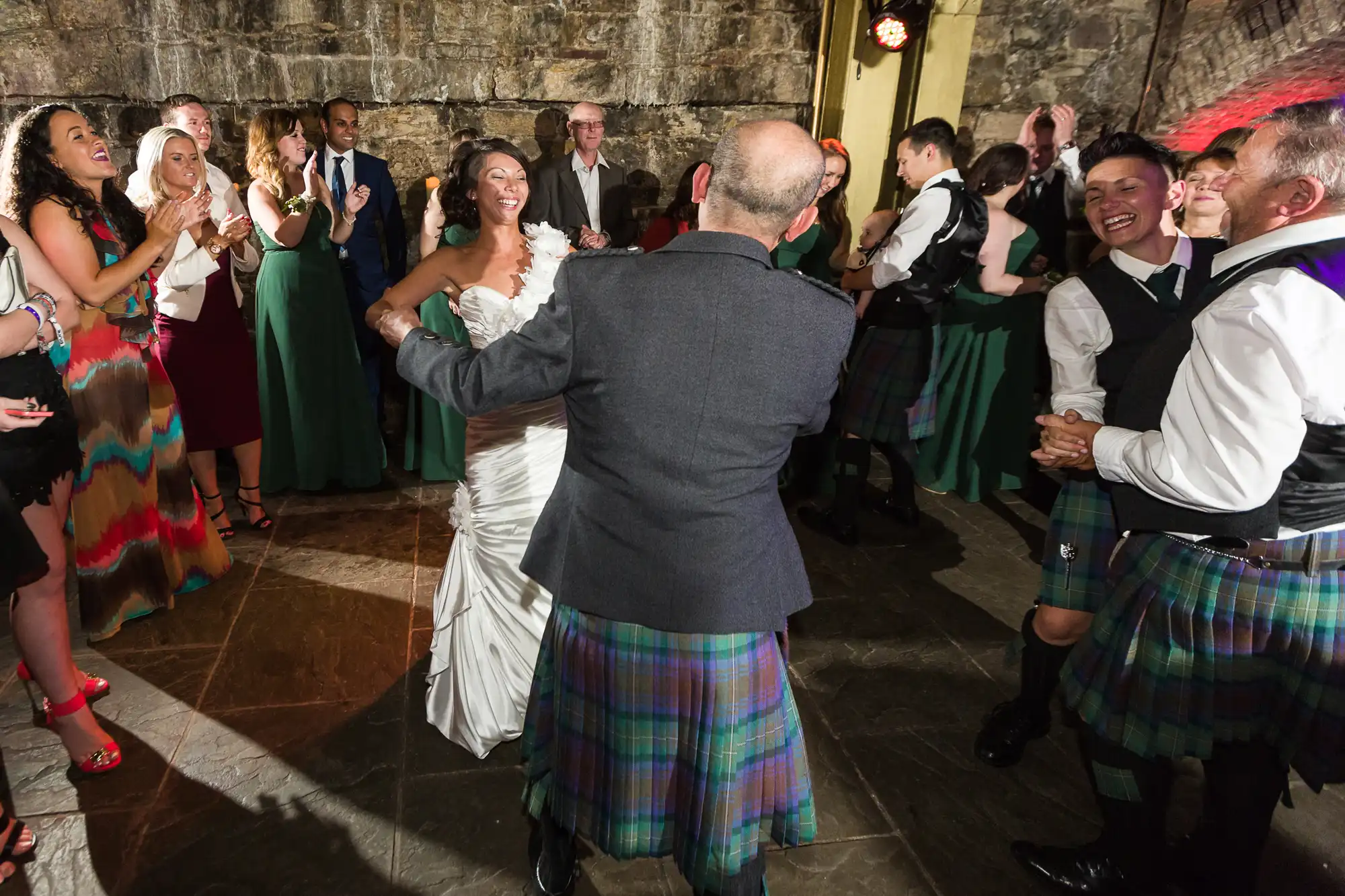 A joyful bride in a white dress dances with a man in a tartan kilt as wedding guests in formal and colourful attire clap and smile around them in a stone-walled venue with warm, festive lighting.