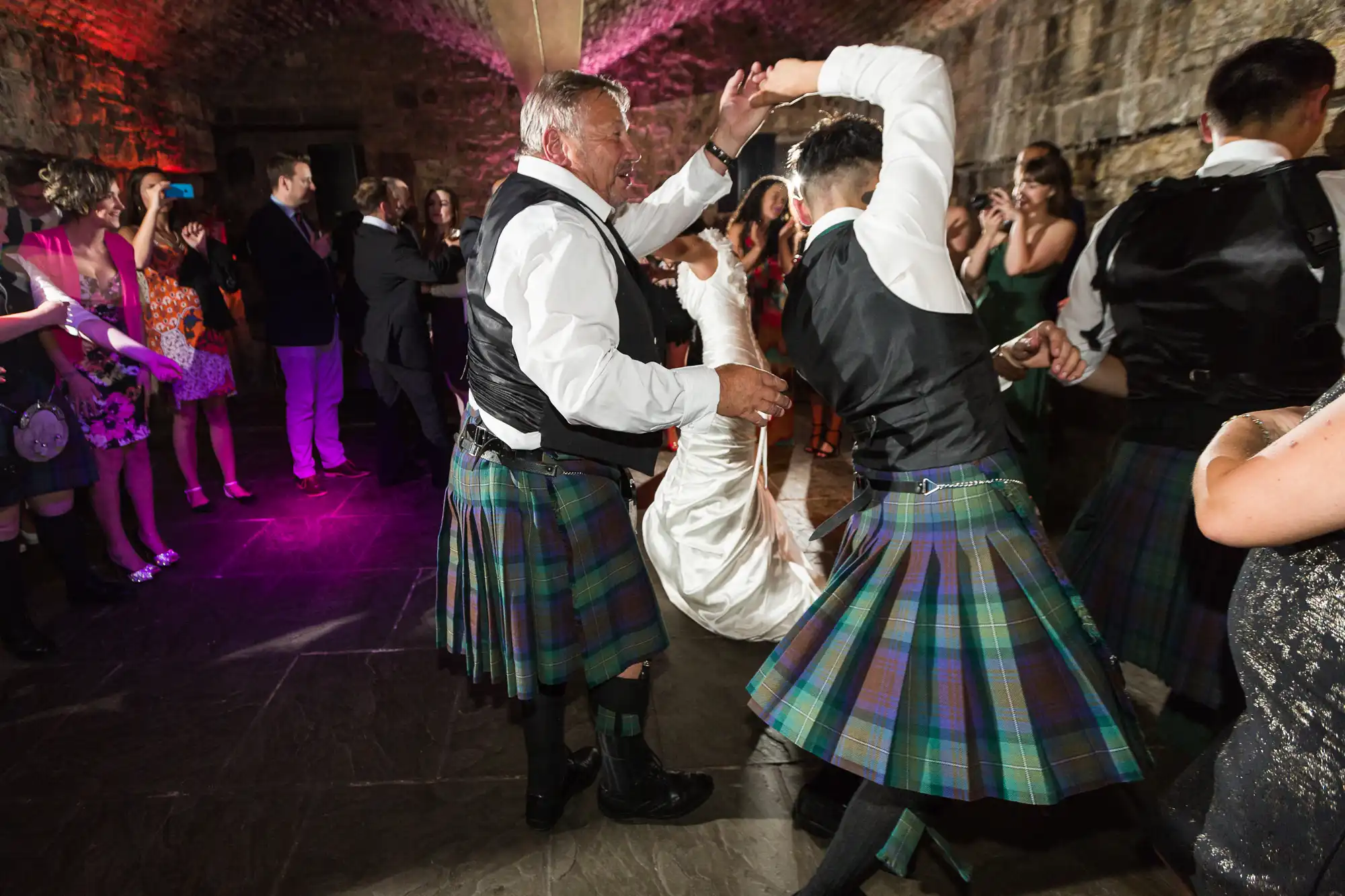 A lively wedding celebration in a stone-walled room shows people dancing energetically. Several men in plaid kilts and waistcoats spin a woman in a white wedding dress. Guests in formal attire watch and smile, and colourful lights brighten the festive scene.