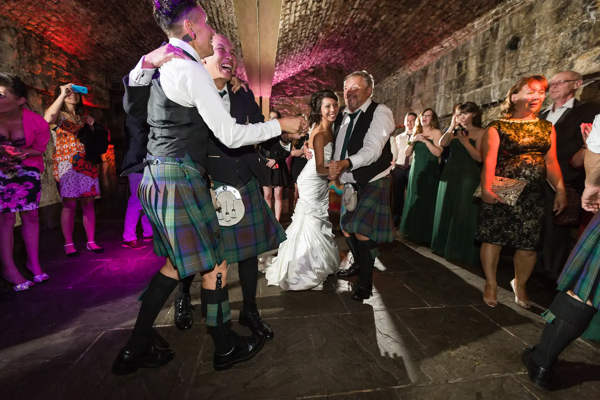 A joyful bride in a white dress dances with men in kilts under a stone-arched ceiling, surrounded by smiling guests. Colourful lighting and festive attire show a lively wedding celebration, with people watching and photographing the moment.