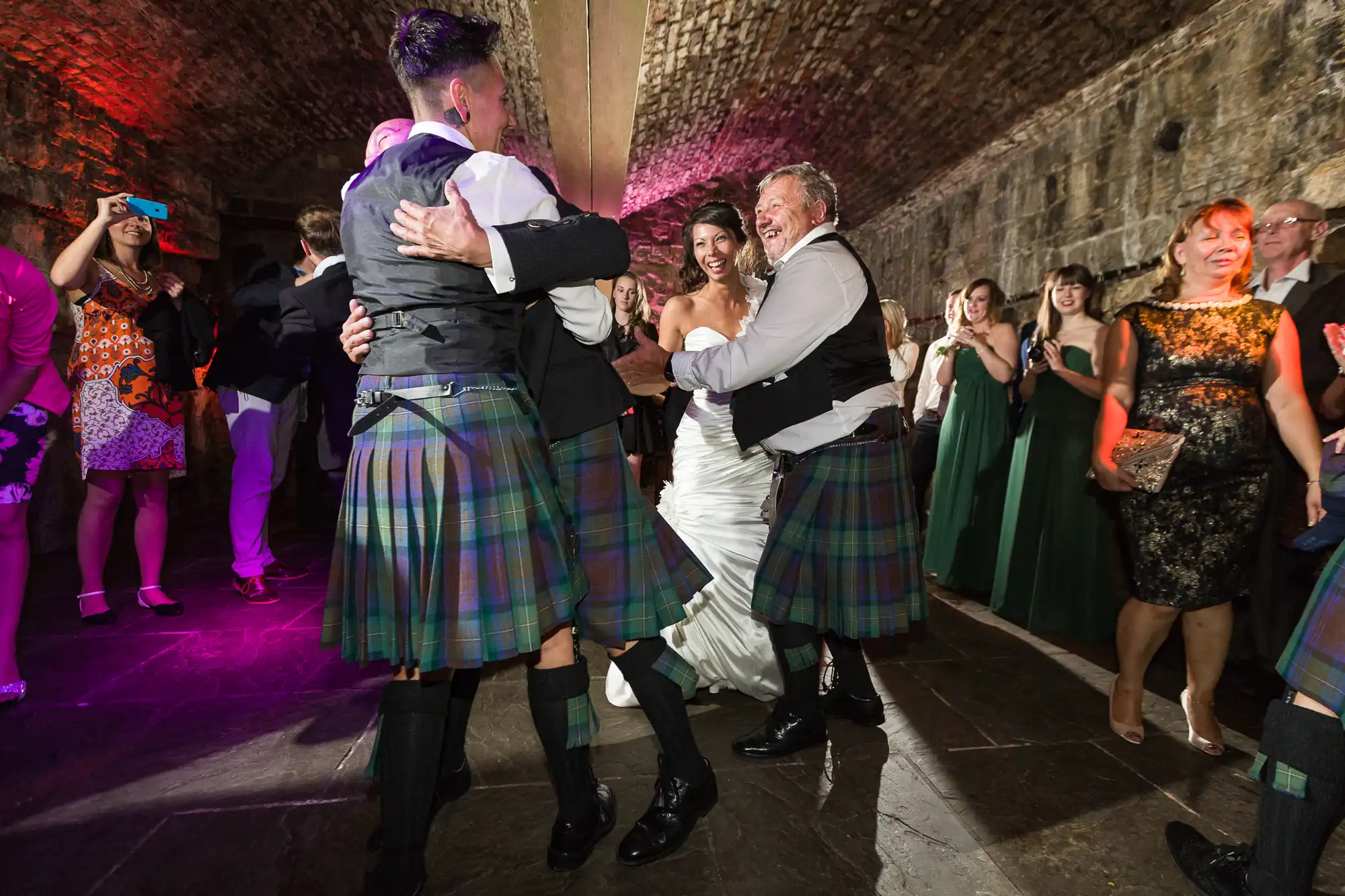 A bride in a white dress dances joyfully with two men in kilts and dress shirts, all smiling and holding hands, surrounded by cheering guests in formal attire under a stone-arched ceiling with colourful lighting.