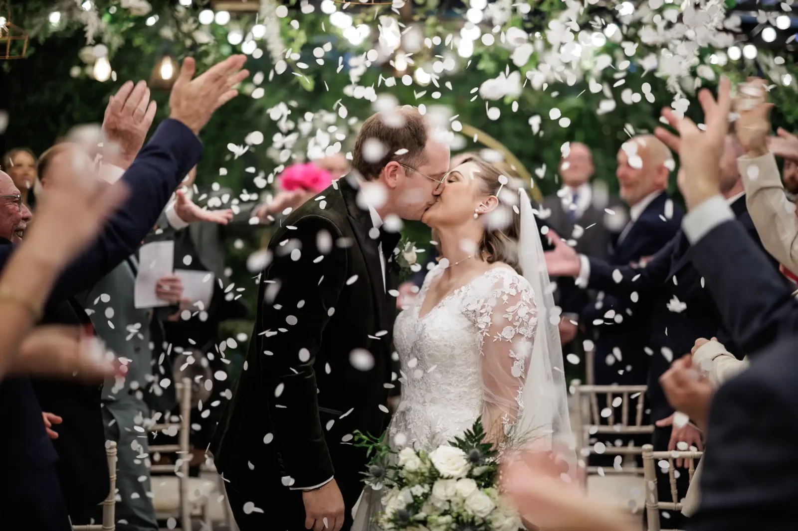 The Scotsman Hotel wedding photos: a bride and groom kiss at their wedding ceremony while guests throw white flower petals in celebration. The bride wears a white lace gown and holds a bouquet; the groom wears a dark suit. People in formal attire clap joyfully in the background.