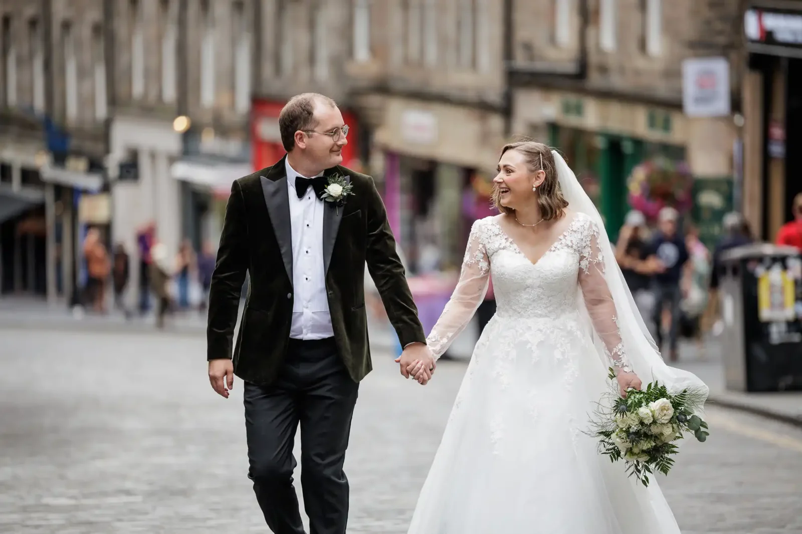 A newlywed couple walk hand in hand on a cobbled street. The bride wears a white dress and veil, holding a bouquet of white flowers, while the groom wears a dark suit with a buttonhole. Both are smiling at each other with buildings in the background.