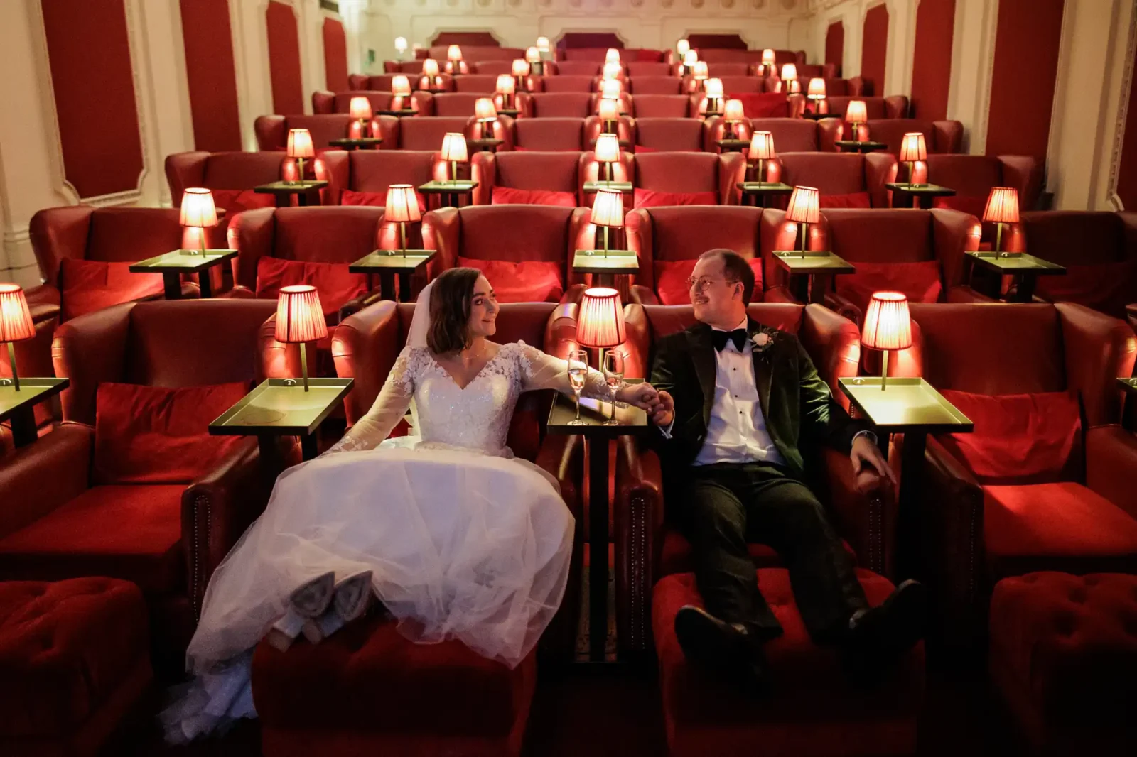 Scotsman Hotel wedding photos: A bride in a white gown and a groom in a tuxedo sit holding hands in plush red theatre seats, surrounded by small lit lamps on tables, smiling at each other in an elegant, empty theatre with rows of red armchairs.