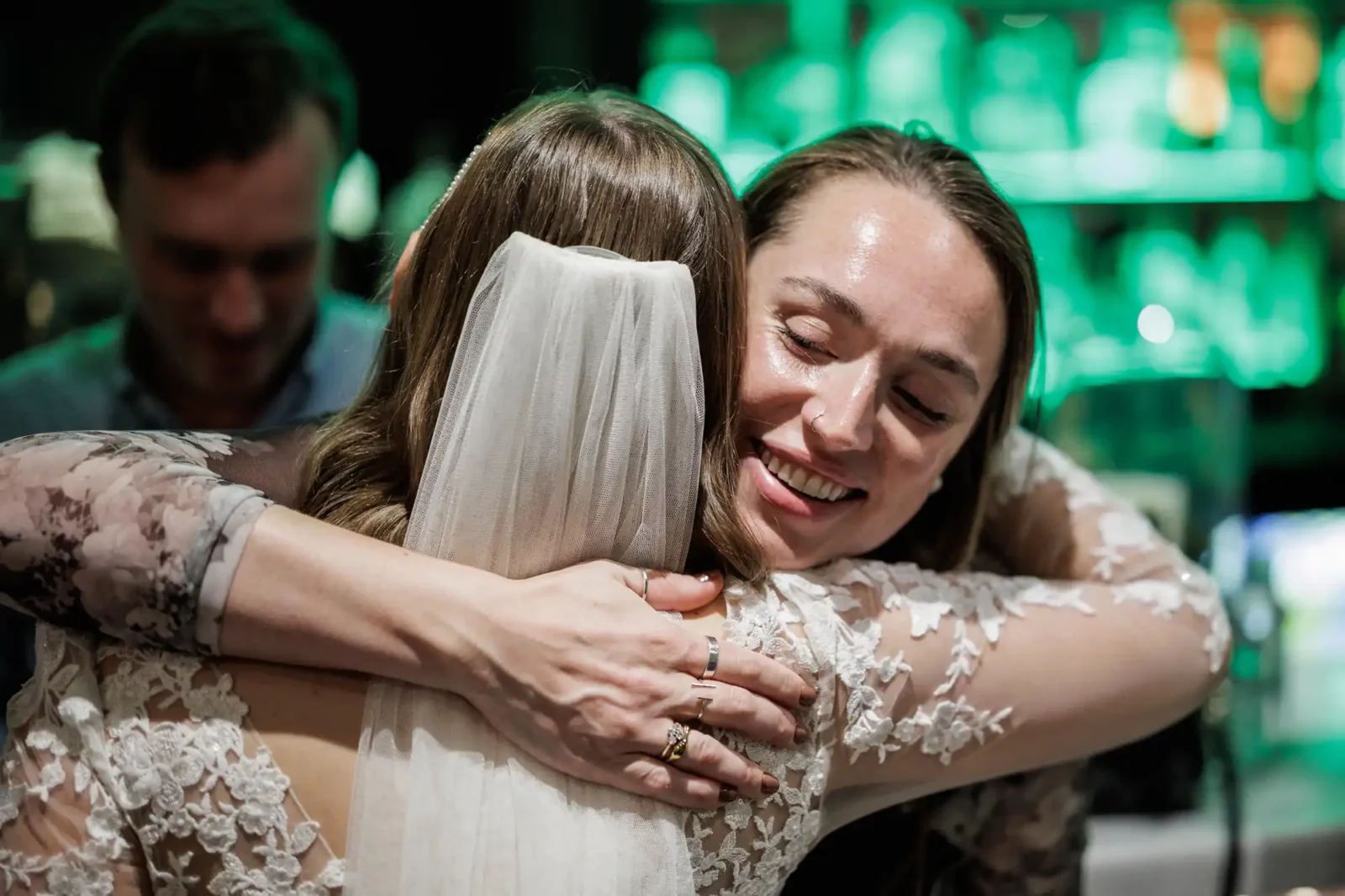 A bride wearing a white lace dress and veil hugs a smiling woman in a similar lace outfit. The woman’s face shows joy as she embraces the bride. Blurred green-lit bar shelves and another person are visible in the background.