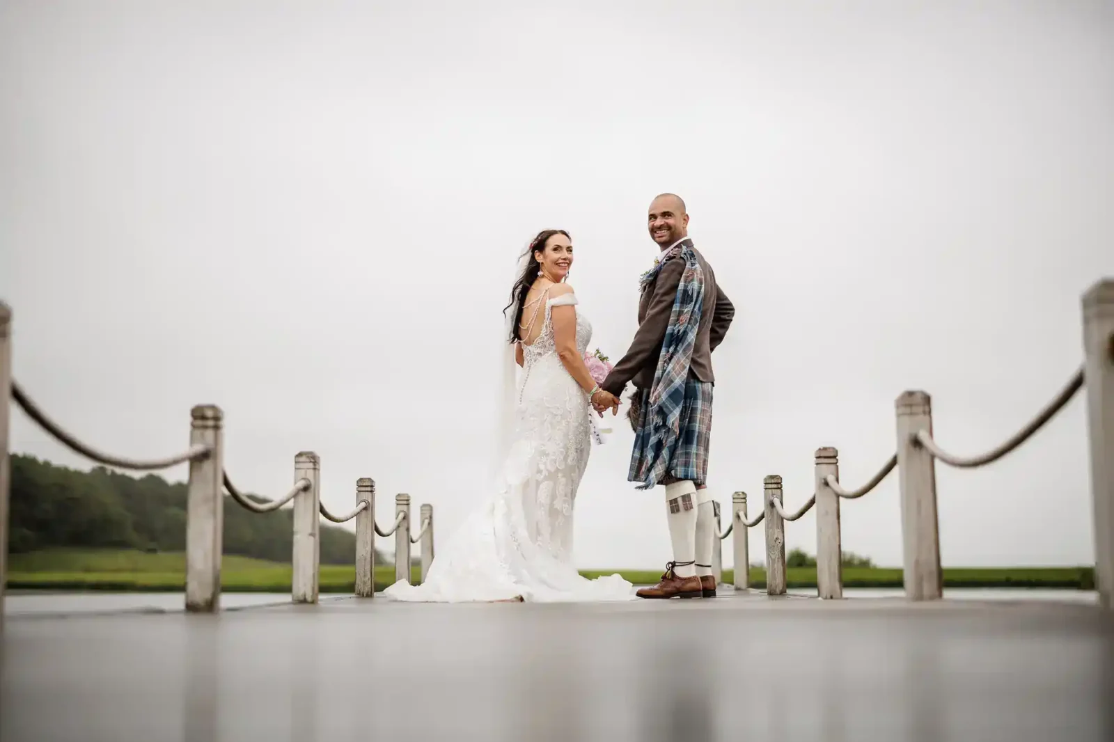 A bride in a white dress and a groom in a kilt stand hand-in-hand on a jetty, smiling back at the camera. The sky is overcast, and greenery is visible in the distance. Ropes line both sides of the jetty.