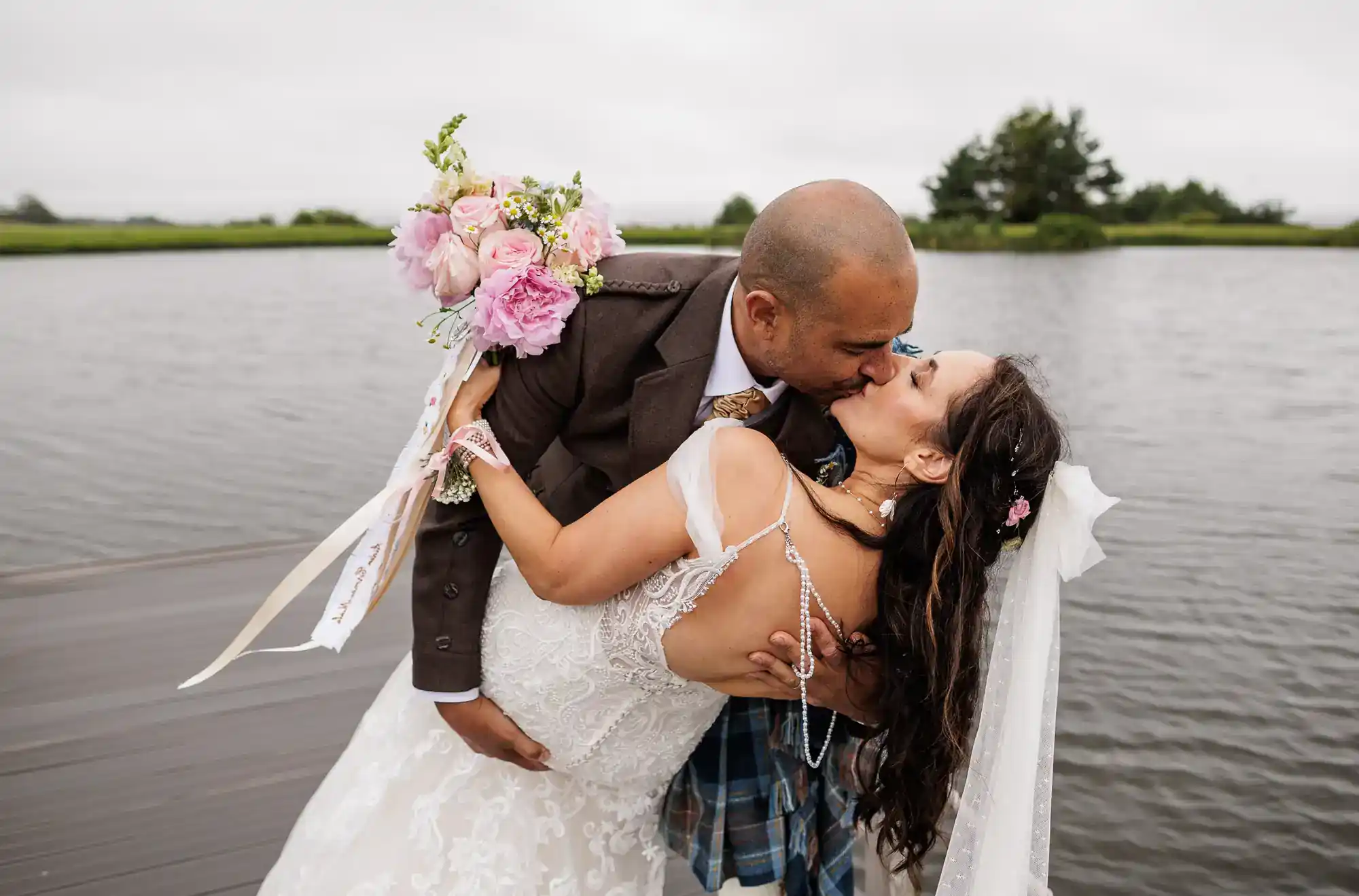 A groom in a brown suit kisses his smiling bride, who holds a pink bouquet and wears a lace dress and veil, as he dips her on a wooden jetty by a lake under an overcast sky. Trees and greenery are visible in the background at The Vu near Bathgate.