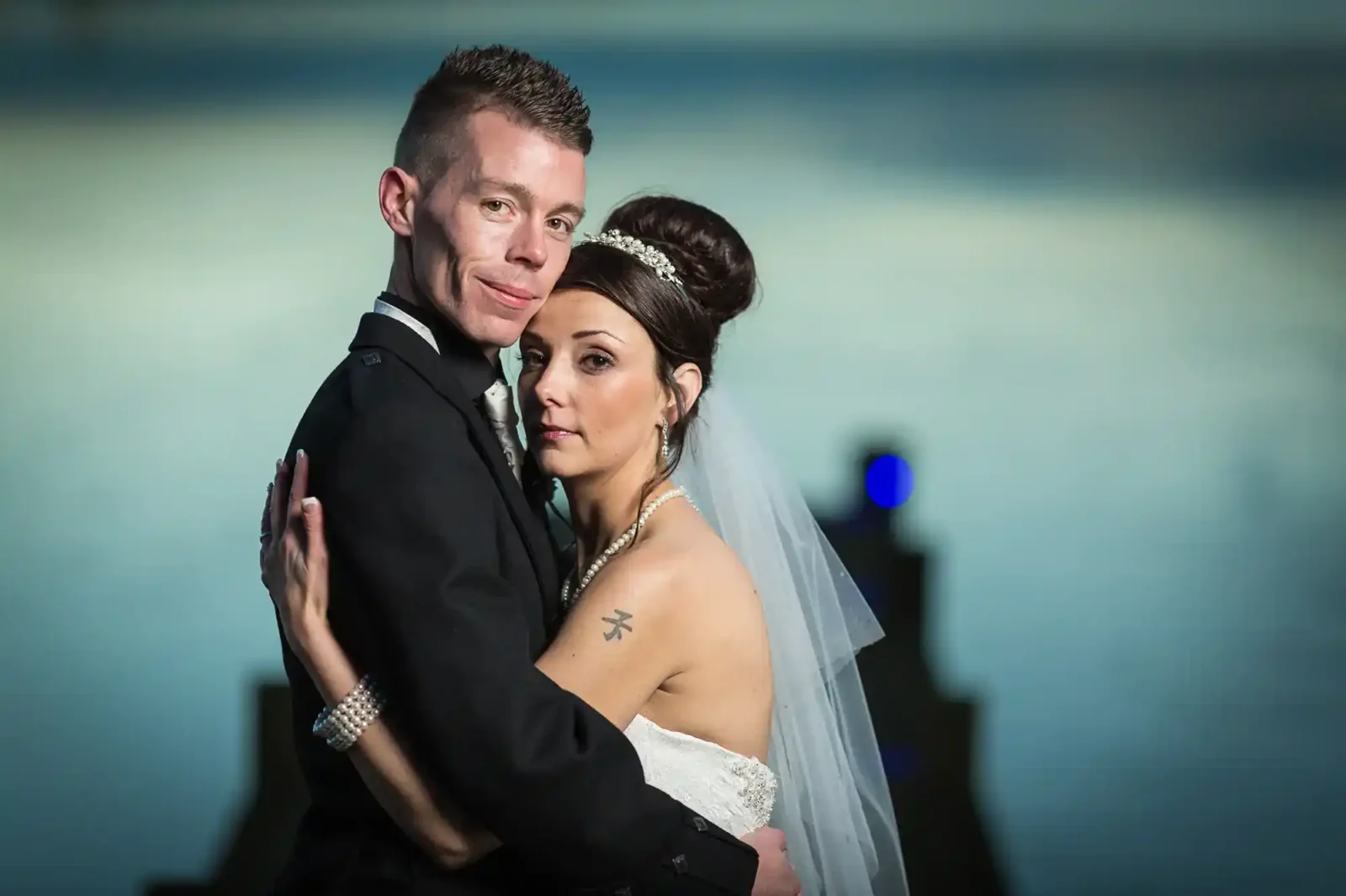 A bride in a strapless white gown with a veil and tiara embraces a groom in a dark suit. They stand close, looking at the camera, against a softly blurred blue background. The bride has a visible tattoo on her shoulder and elegant bridal jewellery.