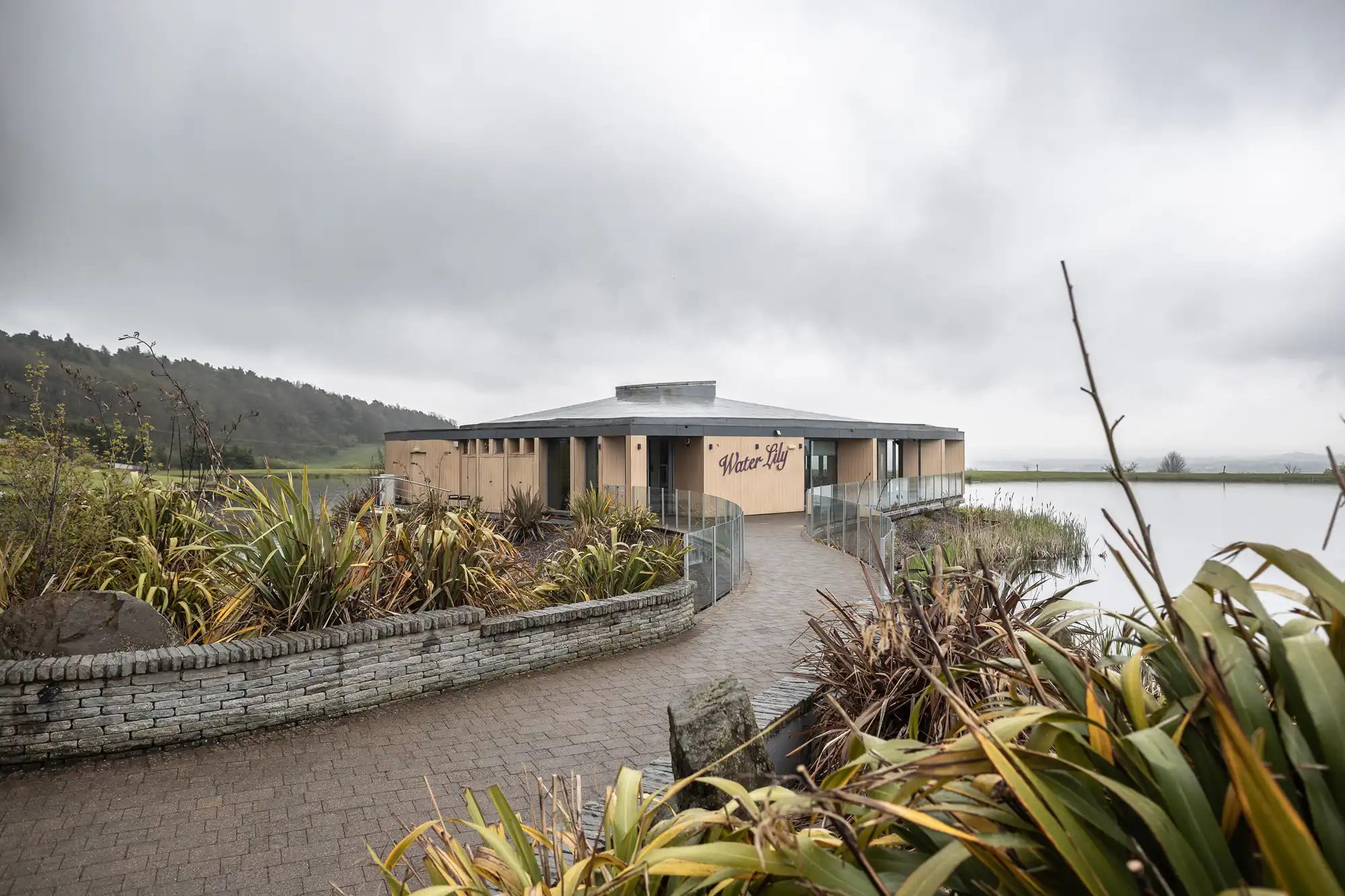 A round, tan-coloured building with the sign "Wharerau" sits by the water, surrounded by greenery and a curved brick pathway. The sky is overcast and misty, and hills are visible in the background.