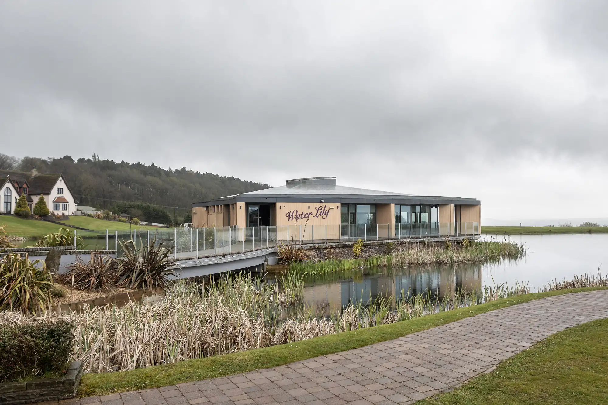 A modern, light-coloured building labelled “Water-Lily” sits beside a pond with reeds and plants. A glass-railed walkway leads to the entrance. Cloudy skies loom overhead, and a path runs along the pond. In the distance, there’s a white house and wooded hills.