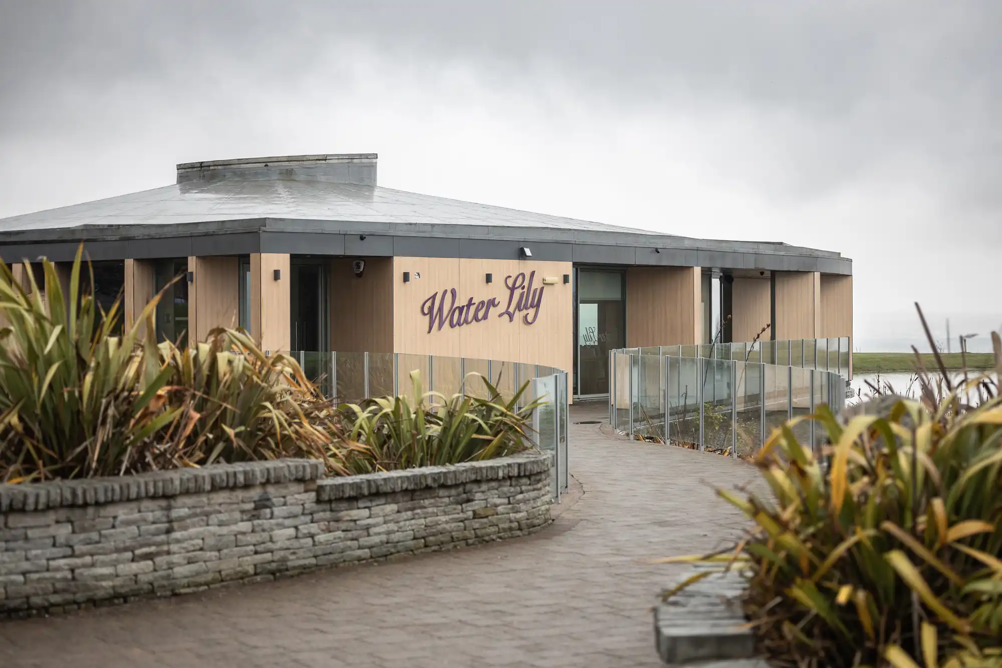 A modern, light-wood building with "Water Lily" written on the side sits behind clear glass fencing. The path is paved with bricks and lined with green plants. Overcast, grey skies and grassy landscape are visible in the background.