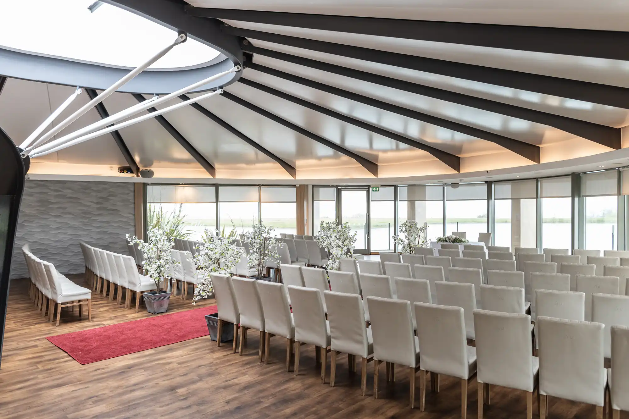 A modern, circular event space with rows of white chairs facing a small altar decorated with white flowers. The room has a wooden floor, large windows with natural light, and a partially open domed ceiling with exposed beams. A red carpet leads to the altar.