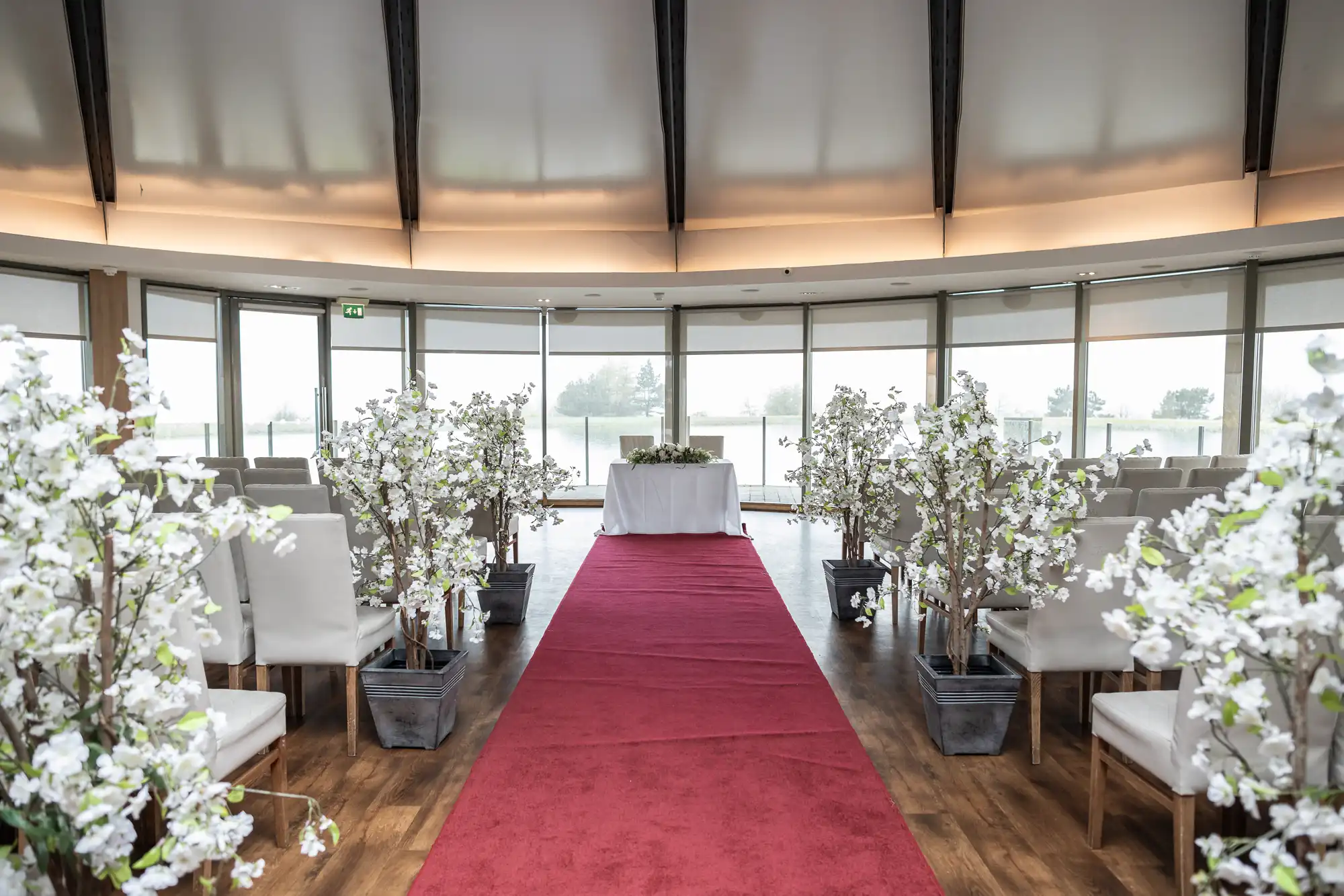 A modern wedding venue with a red carpet aisle lined with potted white blossom trees, surrounded by white chairs. At the end of the aisle is a table with white cloth, set against large, curved windows letting in natural light.