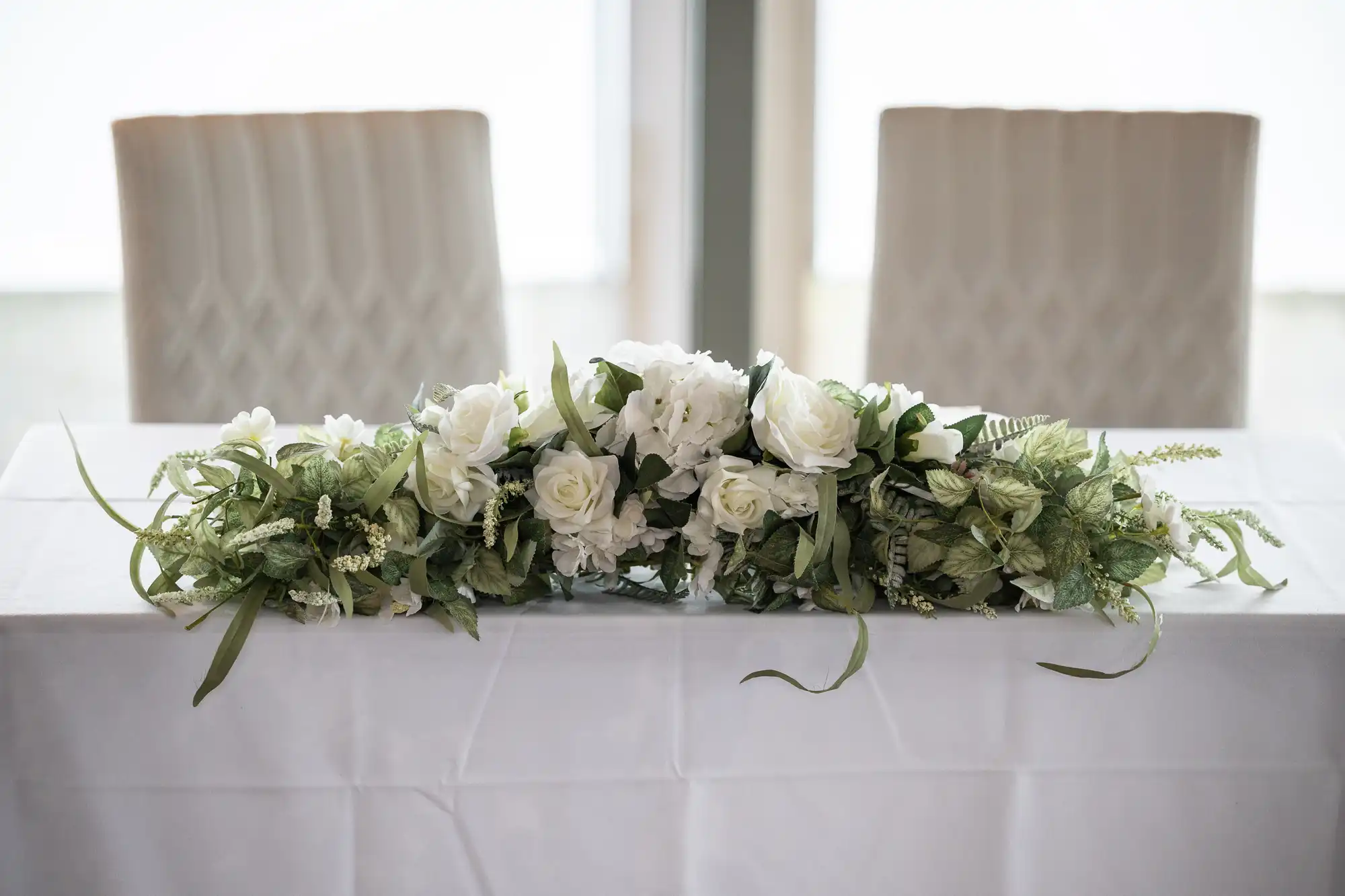 A white tablecloth covers a table topped with a floral arrangement of white roses and greenery. Behind the table, two empty, cream-coloured chairs with a textured pattern are visible, and a softly lit window is in the background.