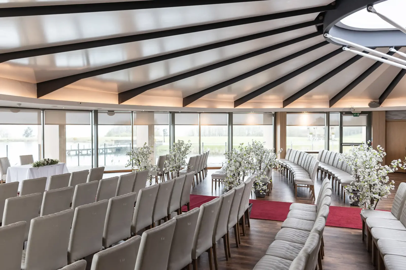 A bright, modern wedding venue with rows of cream chairs facing a small table. Large windows overlook a green landscape. White flower arrangements line the aisle, and red carpets create two curved pathways down the centre of the wooden floor.