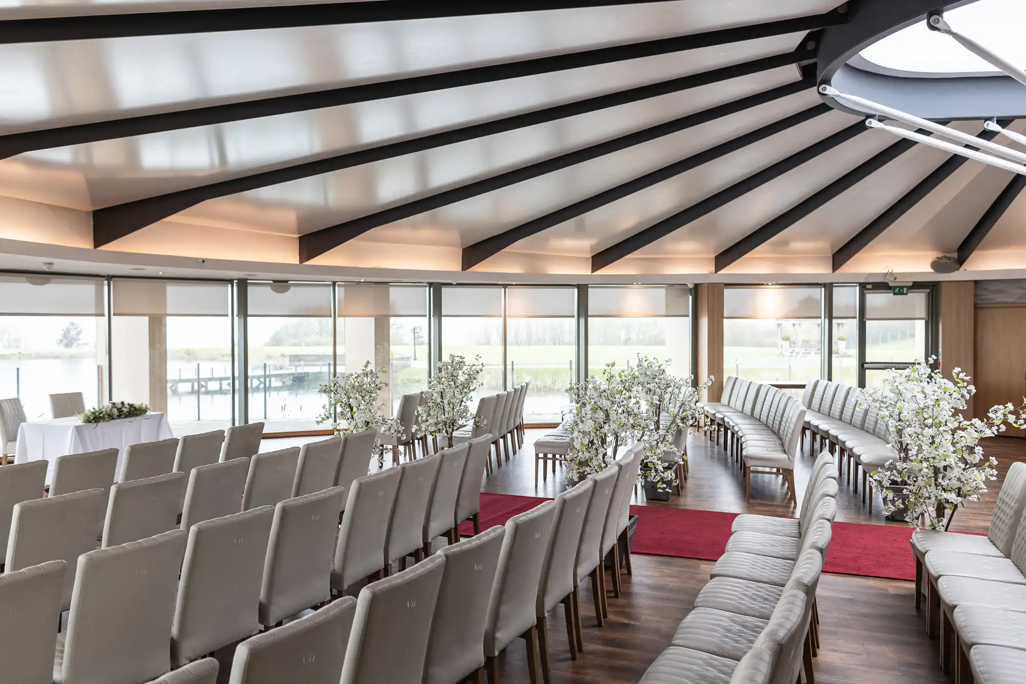 A bright, modern wedding venue with rows of cream chairs facing a small table. Large windows overlook a green landscape. White flower arrangements line the aisle, and red carpets create two curved pathways down the centre of the wooden floor.