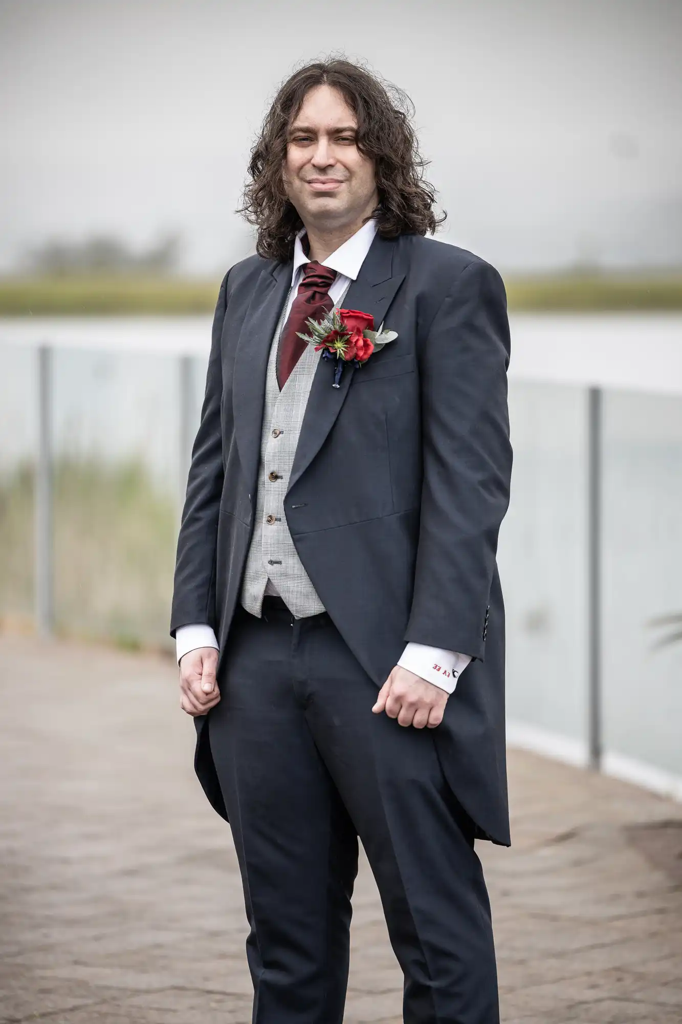 A man with long curly brown hair stands outdoors on a wooden path by a glass balustrade. He wears a dark suit with a grey waistcoat, white shirt, maroon cravat, and buttonhole with red flowers. The background is blurred with greenery and water.