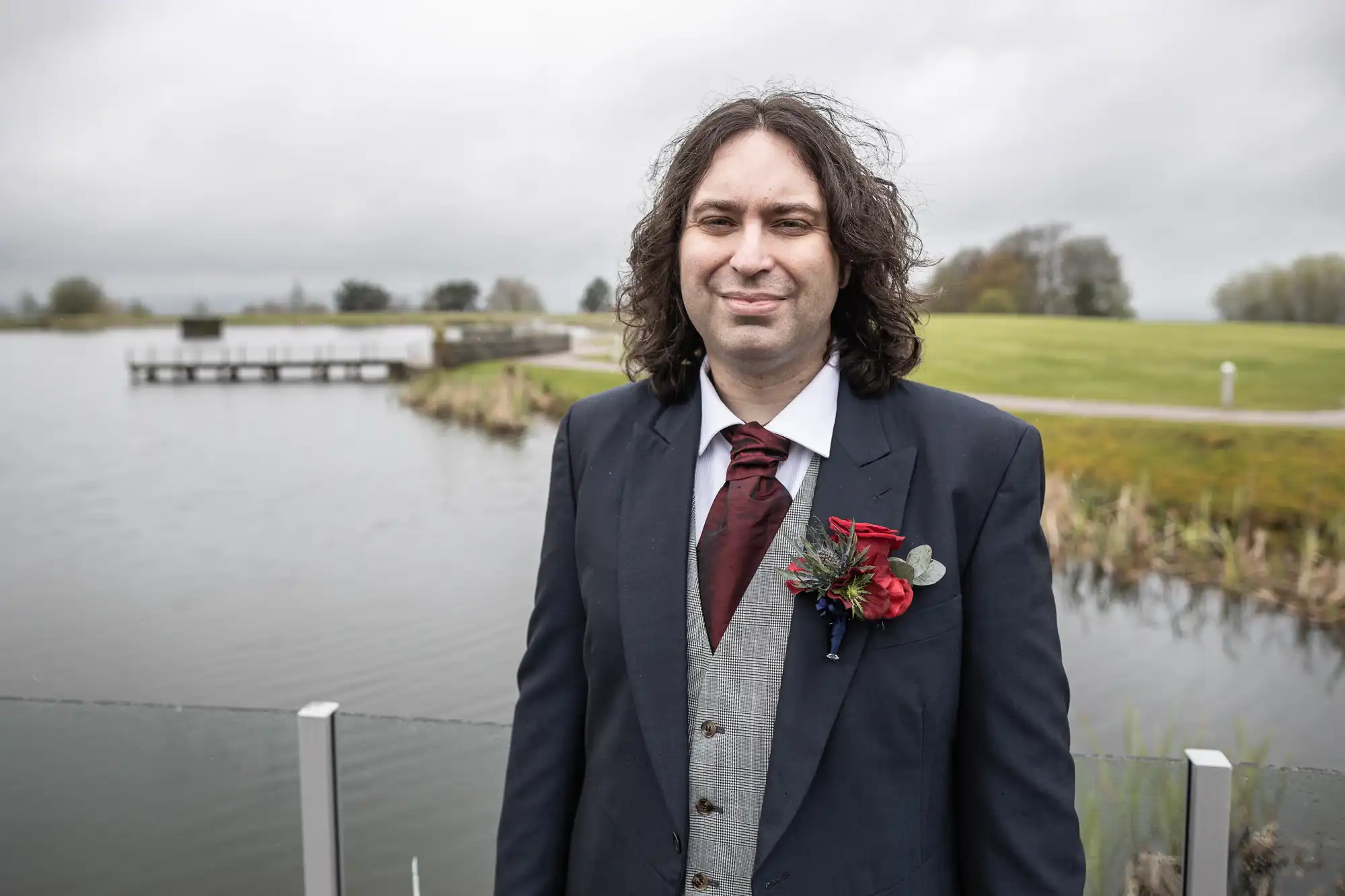 A man with long curly hair, wearing a dark suit, checked waistcoat, white shirt, and burgundy tie, stands by a lake. He has a red buttonhole on his lapel. The background shows grass, water, a jetty, and cloudy sky.