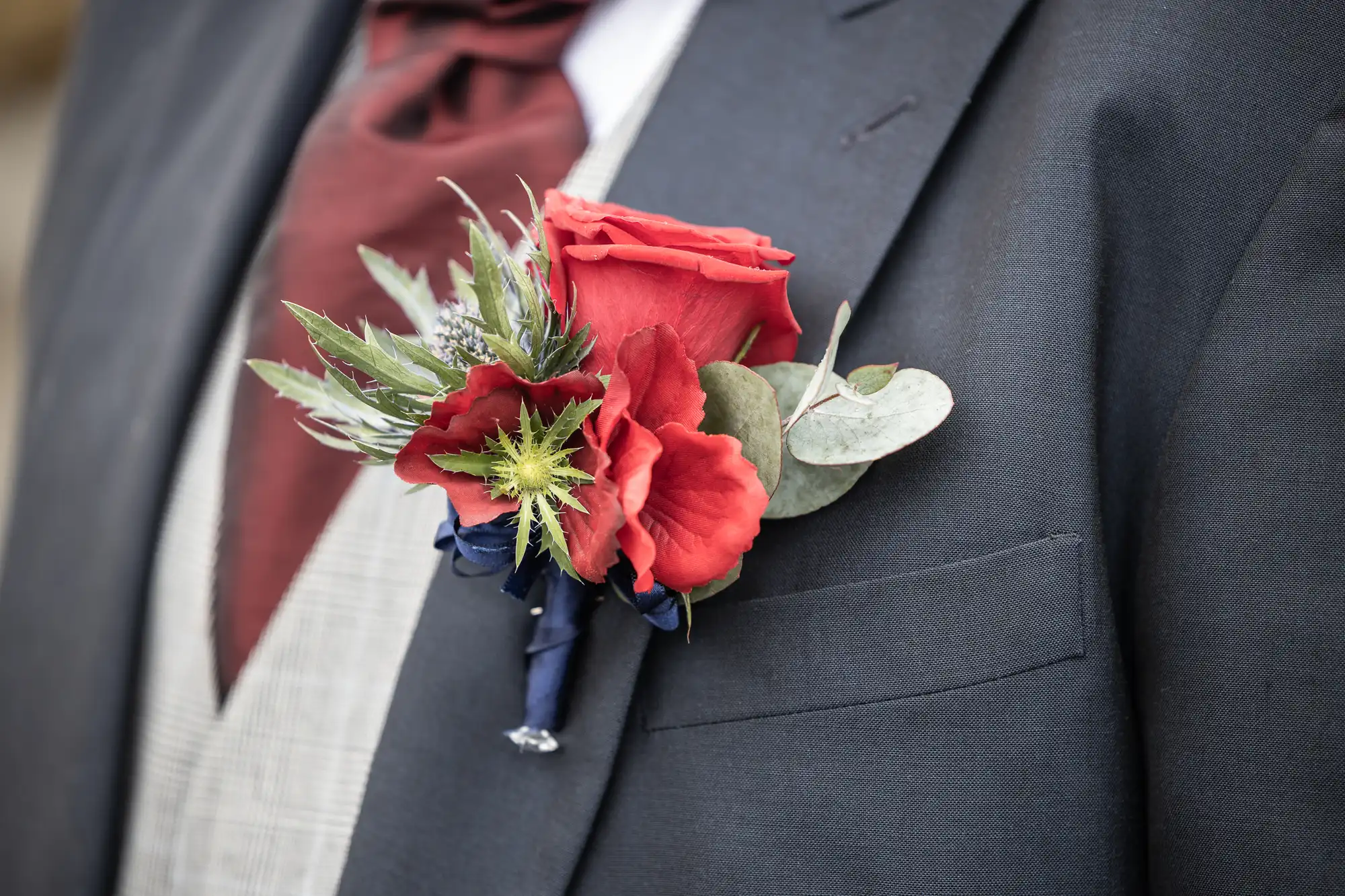 A close-up of a person wearing a dark grey suit with a red rose buttonhole pinned to the lapel. The buttonhole also includes green leaves and thistles, and the person has a maroon cravat and a light check waistcoat underneath the suit jacket.