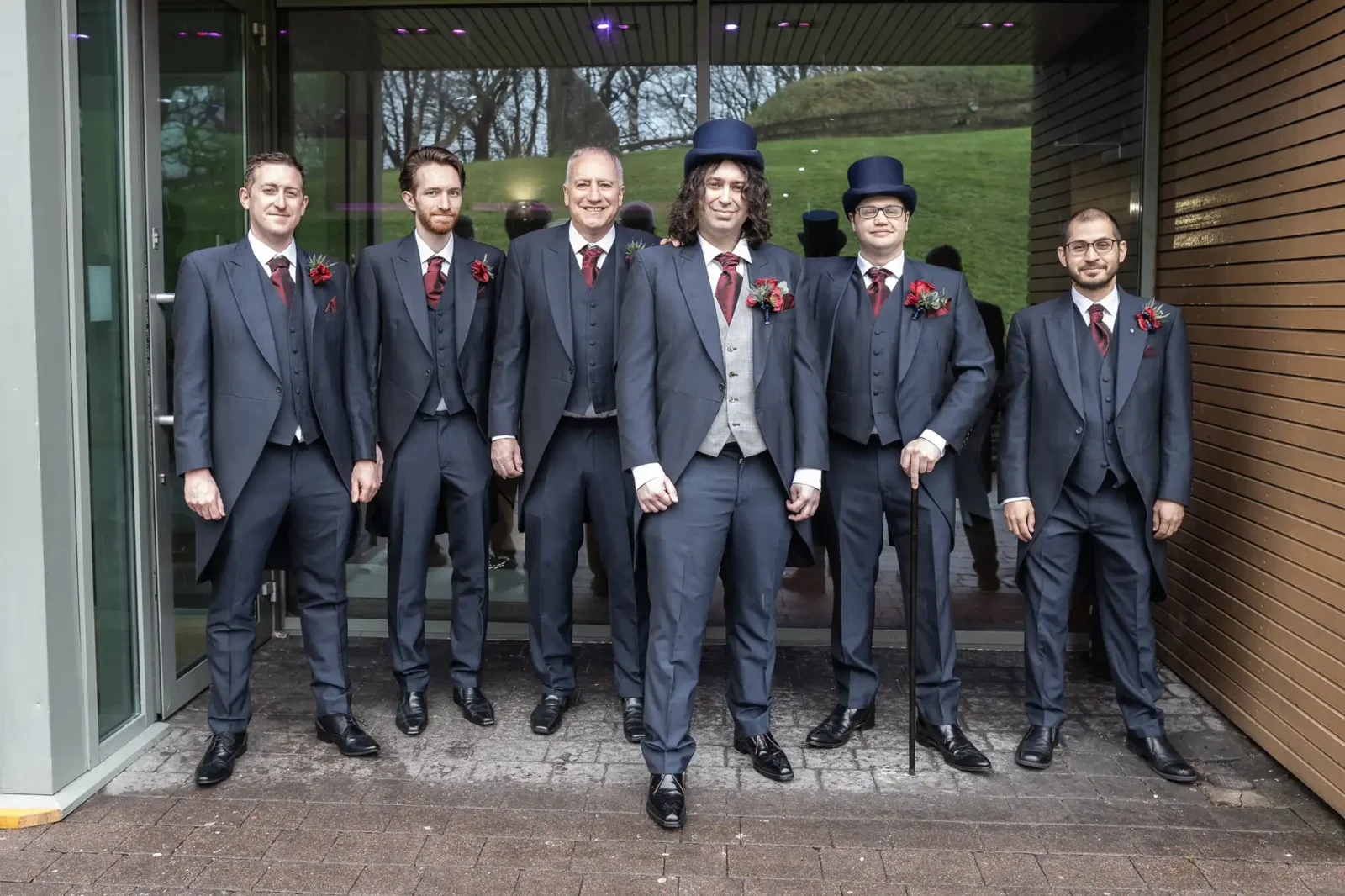 The Vu wedding photos of groom John, with five groomsmen in matching dark suits, white shirts, and red buttonholes stand outside a building. Two wear top hats; one holds a cane. They smile and pose together in a row. Glass doors and a wooden wall are behind them, with greenery visible outside.