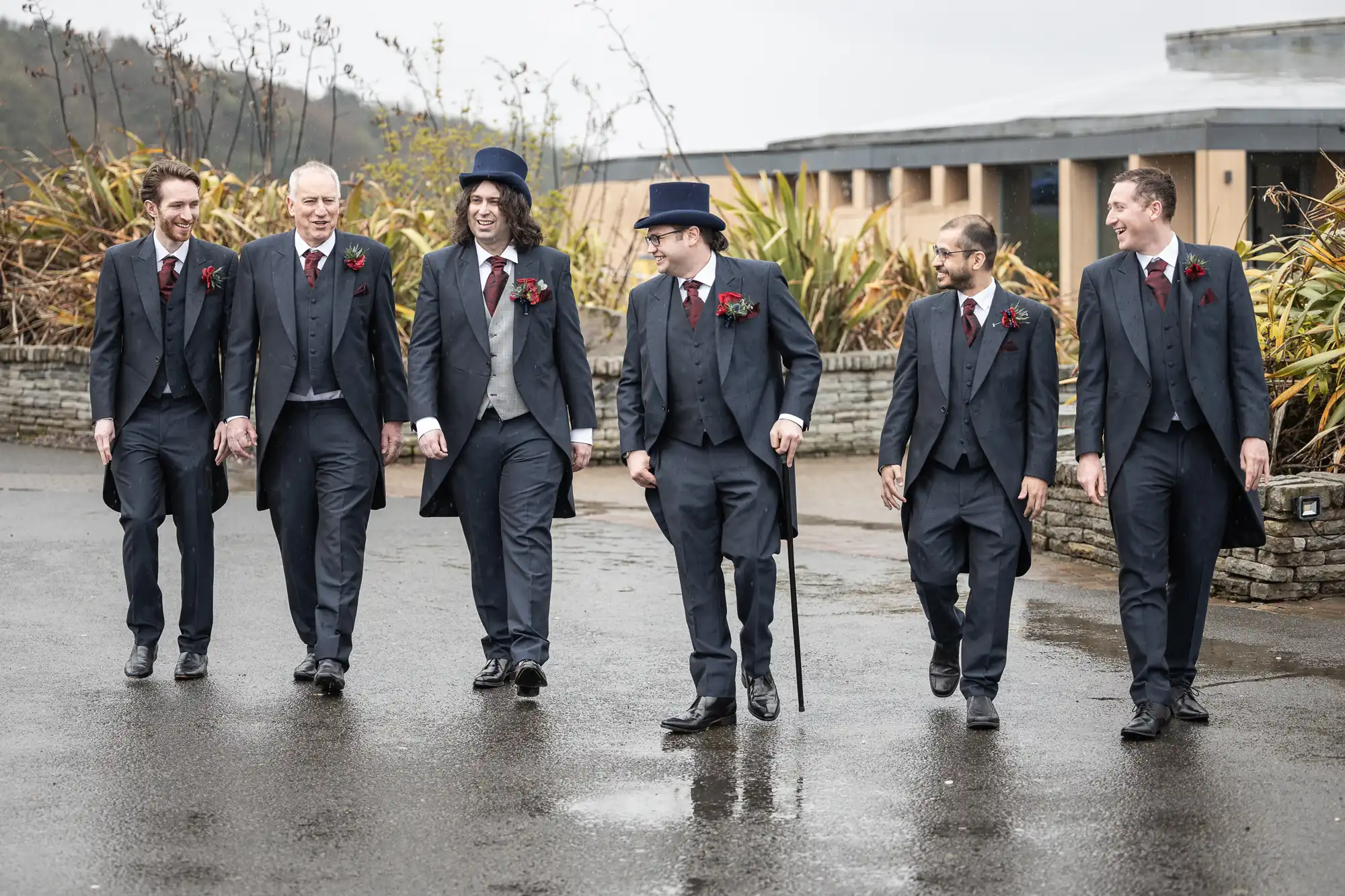 Six men in matching dark suits with burgundy ties and buttonholes walk together outdoors on a wet pavement. Two wear top hats, one holds a cane. They are smiling and talking, with plants and a building in the background. The scene looks candid and joyful.