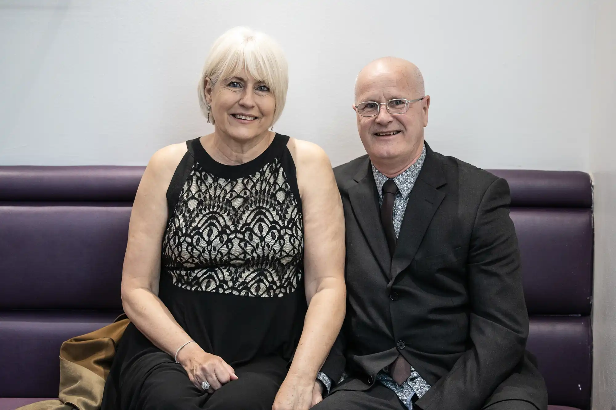 A woman with short, white-blonde hair in a black lace dress sits beside a man with glasses and a suit on a purple bench. Both are smiling at the camera against a plain white wall. The woman's gold handbag rests on the seat beside her.