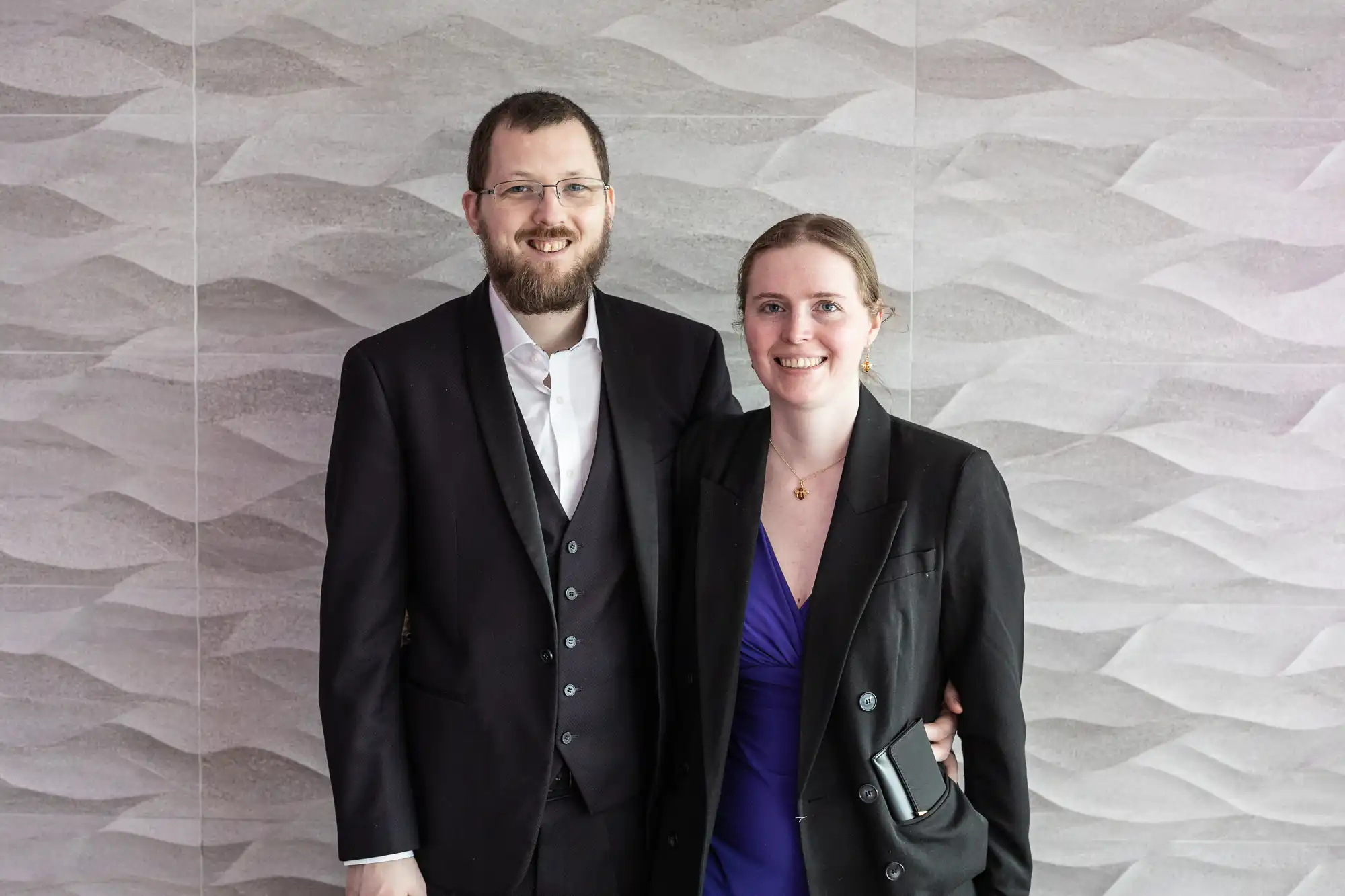 A smiling man and woman stand arm-in-arm in front of a wavy-patterned light grey wall. The man has a beard, glasses, and wears a dark suit with a white shirt. The woman wears a black blazer over a deep purple dress and holds a black clutch.