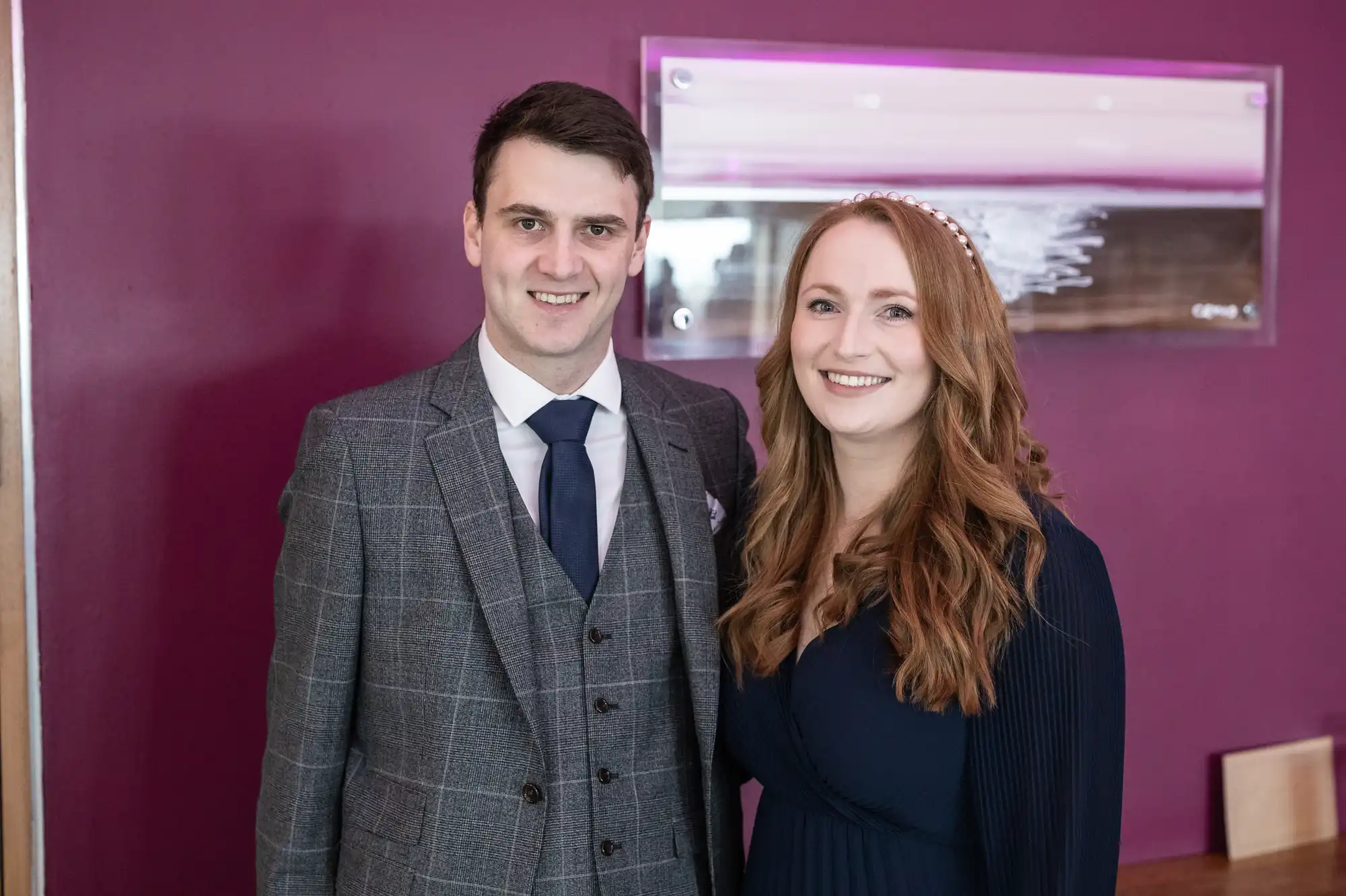 A man in a grey checked suit and navy tie stands next to a woman with long wavy auburn hair, wearing a navy dress and pearl headband. They are smiling in front of a purple wall with a modern, abstract artwork.