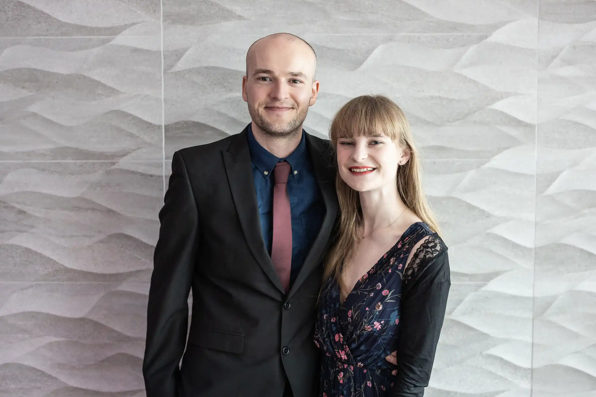 A smiling man in a black suit and red tie stands with his arm round a smiling woman in a navy floral dress with lace sleeves. They pose together in front of a light grey, wavy-textured wall.