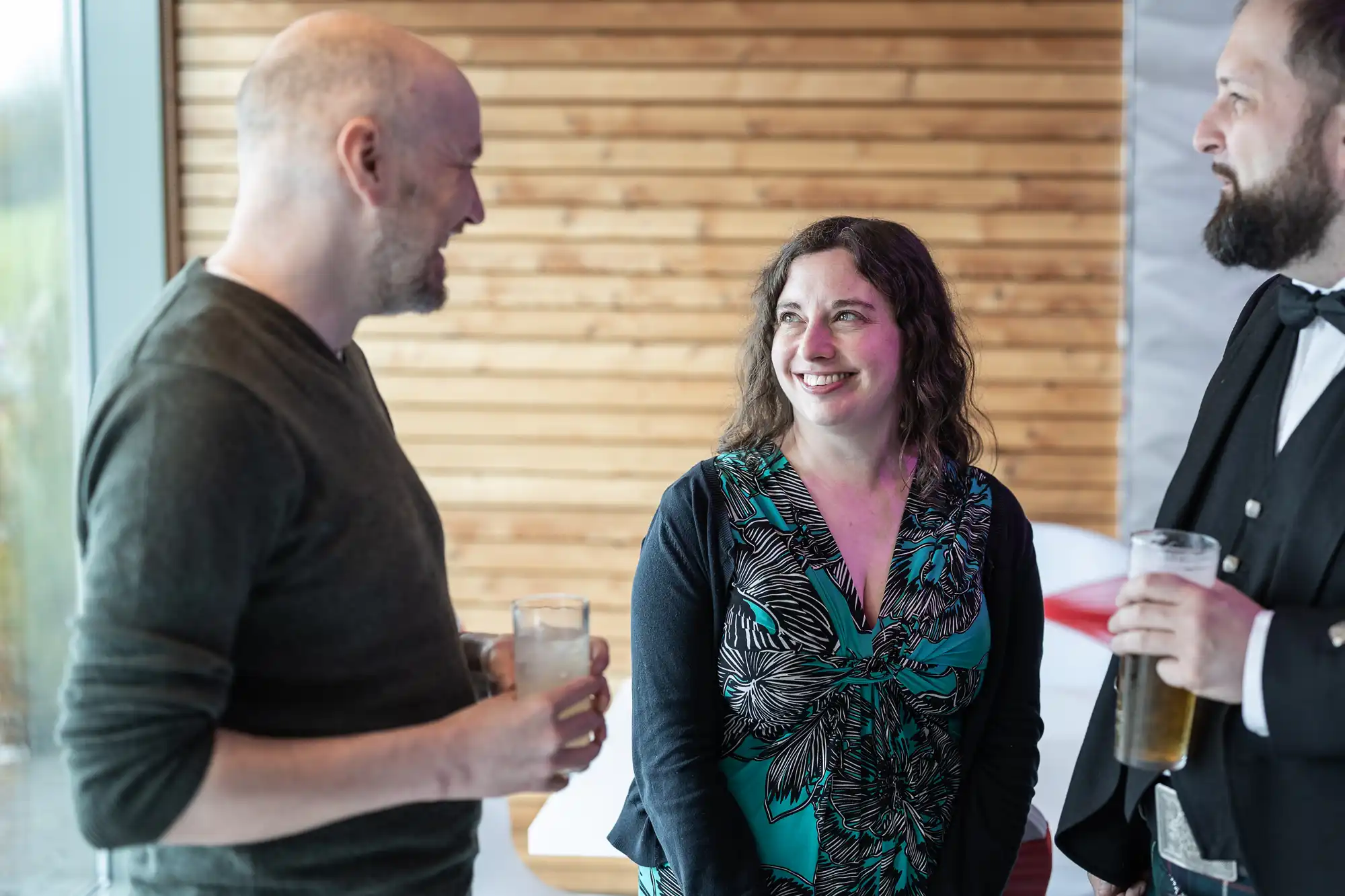 Three adults stand together indoors, smiling and chatting. One man on the left holds a drink, the woman in the centre smiles at him, and a bearded man on the right wears a suit and holds a glass of beer. The background features horizontal wooden panelling.