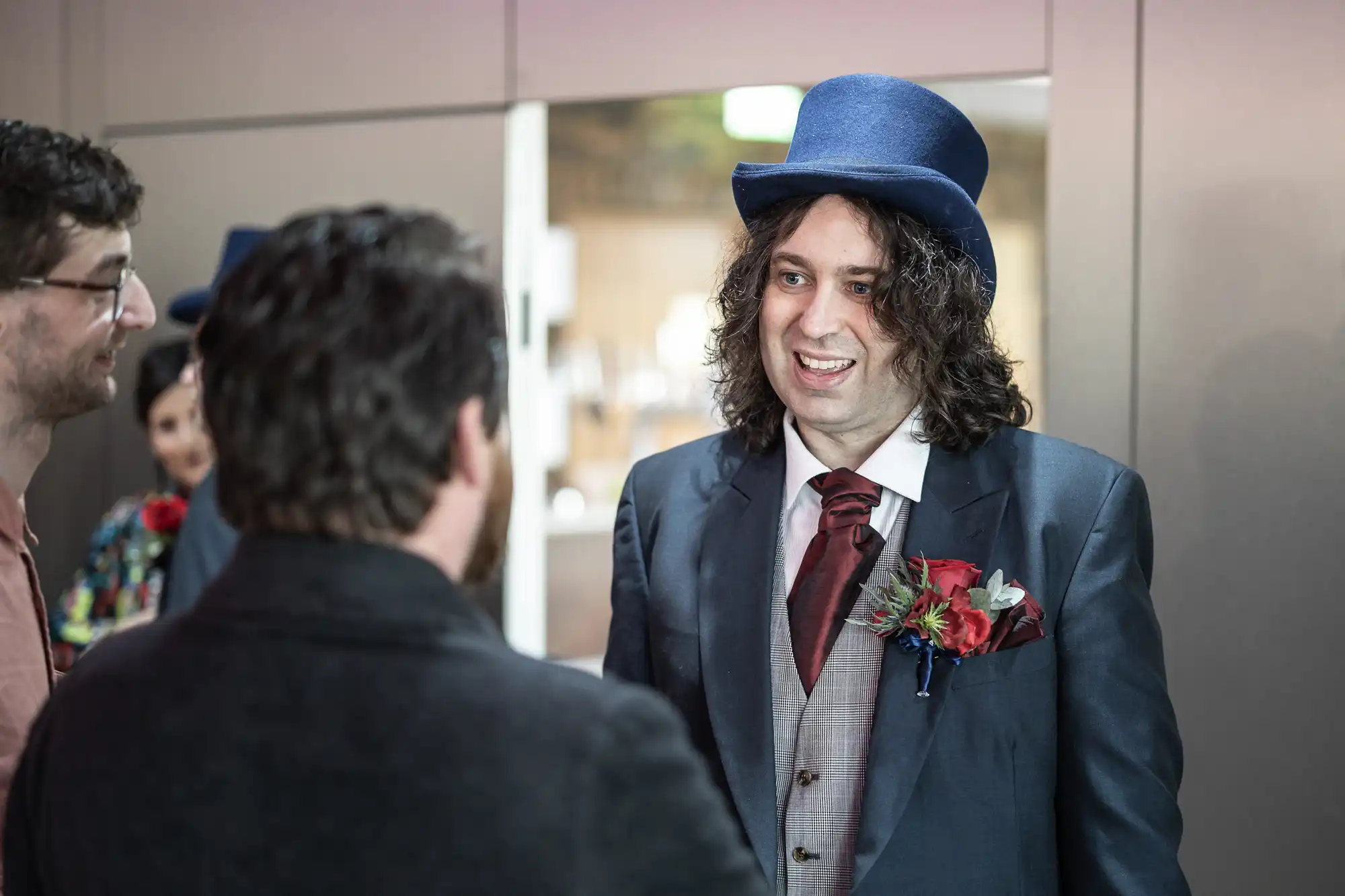 A man with long curly hair wearing a blue top hat, dark suit, and red tie with a colourful buttonhole smiles and talks to two people at an indoor event. Blurred people and a modern, grey background are visible.