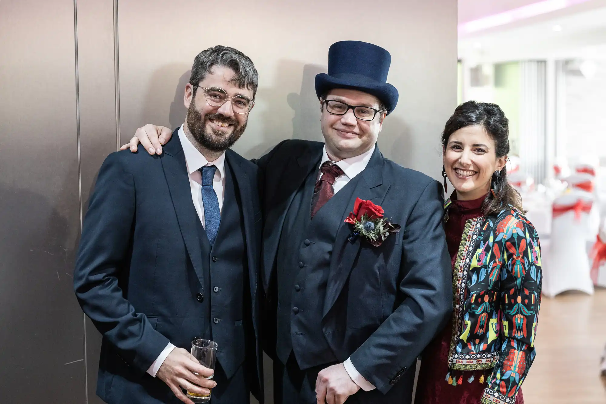 Three adults stand together smiling. The man in the centre wears a dark suit, burgundy tie, buttonhole, and a tall blue hat. The man on the left wears a suit and holds a drink. The woman on the right wears a patterned jacket over a red dress.