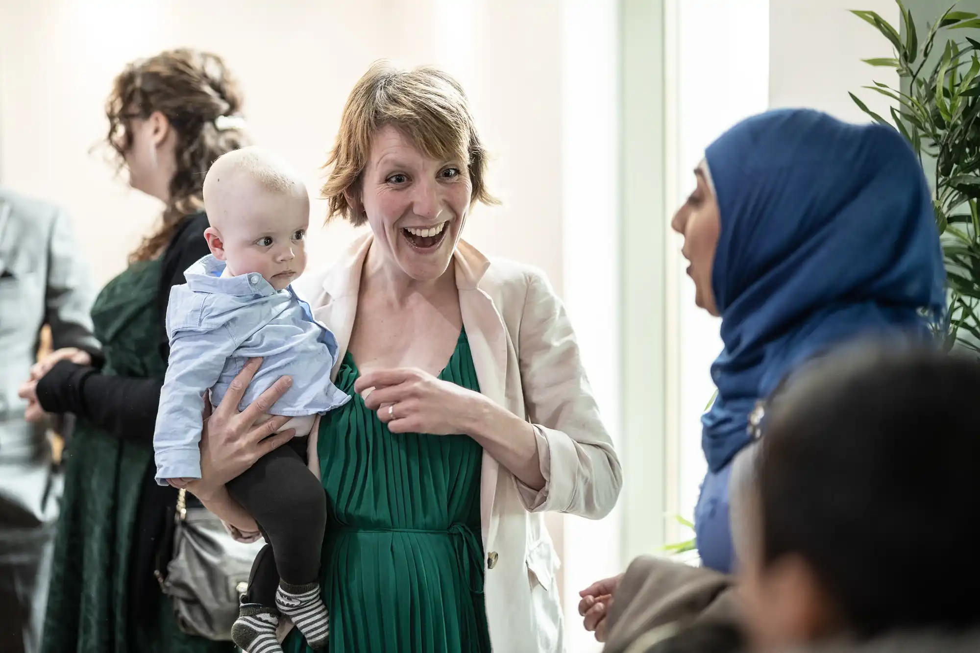 A smiling woman in a light jacket holds a baby who wears a blue shirt. She faces a woman in a blue hijab, speaking animatedly. Other people and green plants are in the background in a bright indoor setting.