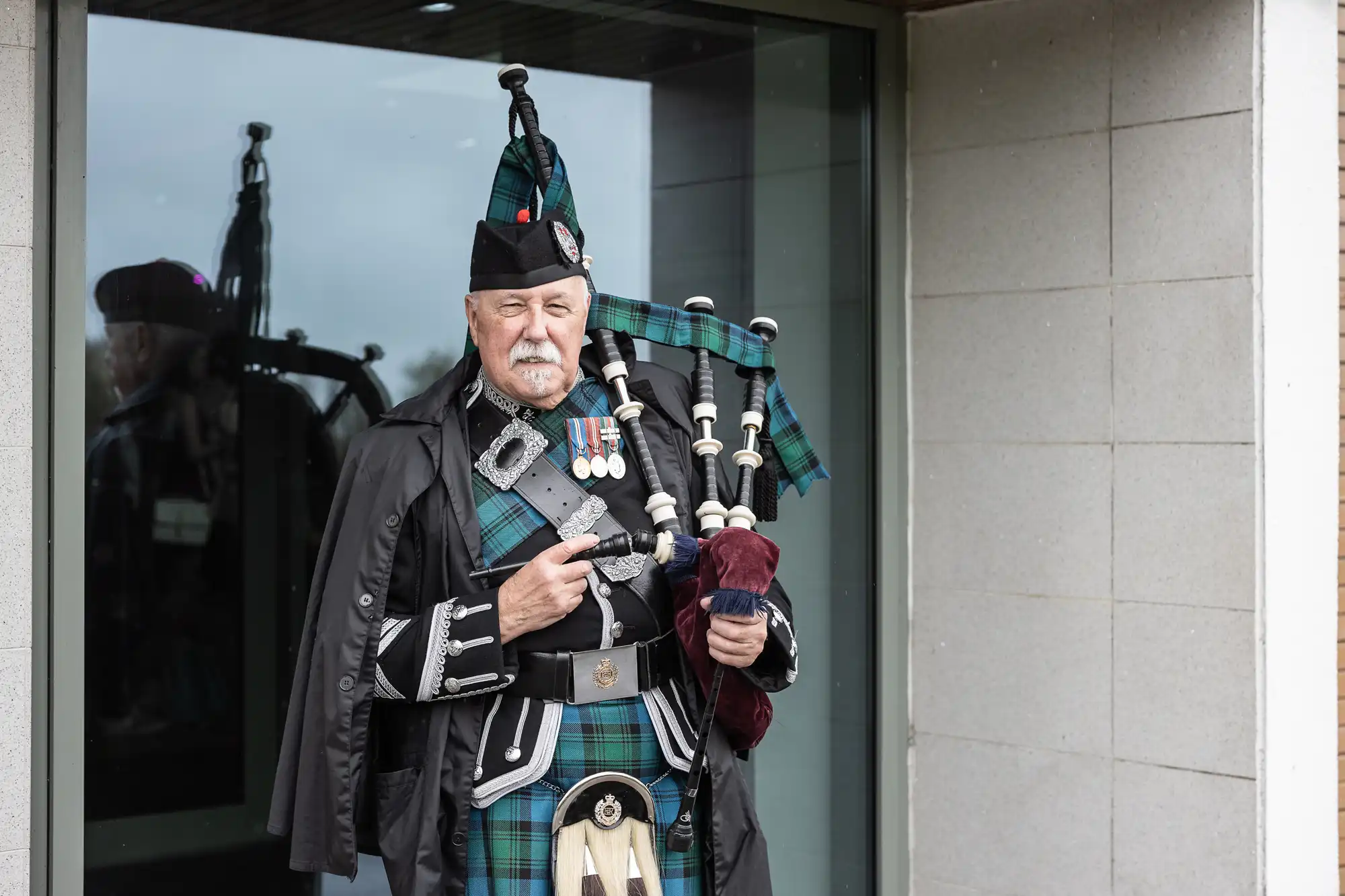 An older man in traditional Scottish attire, including a tartan kilt and bonnet, stands playing the bagpipes. He wears a black cape and several medals on his chest. He stands outside near a glass window and a grey stone wall.