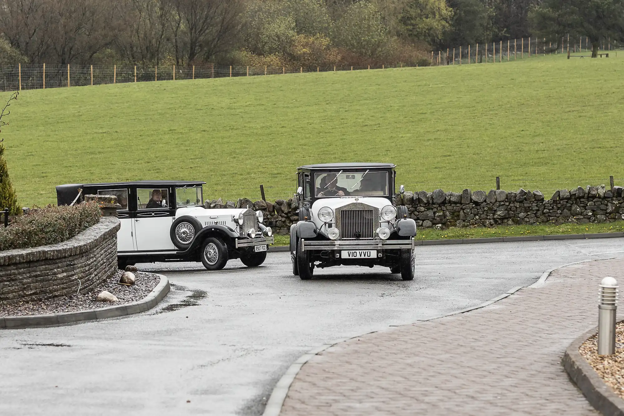 Two vintage cars, one white and one black, drive on a wet road bordered by stone walls and green grassy fields. Trees and a wire fence are visible in the background under an overcast sky. The scene appears rural and peaceful.