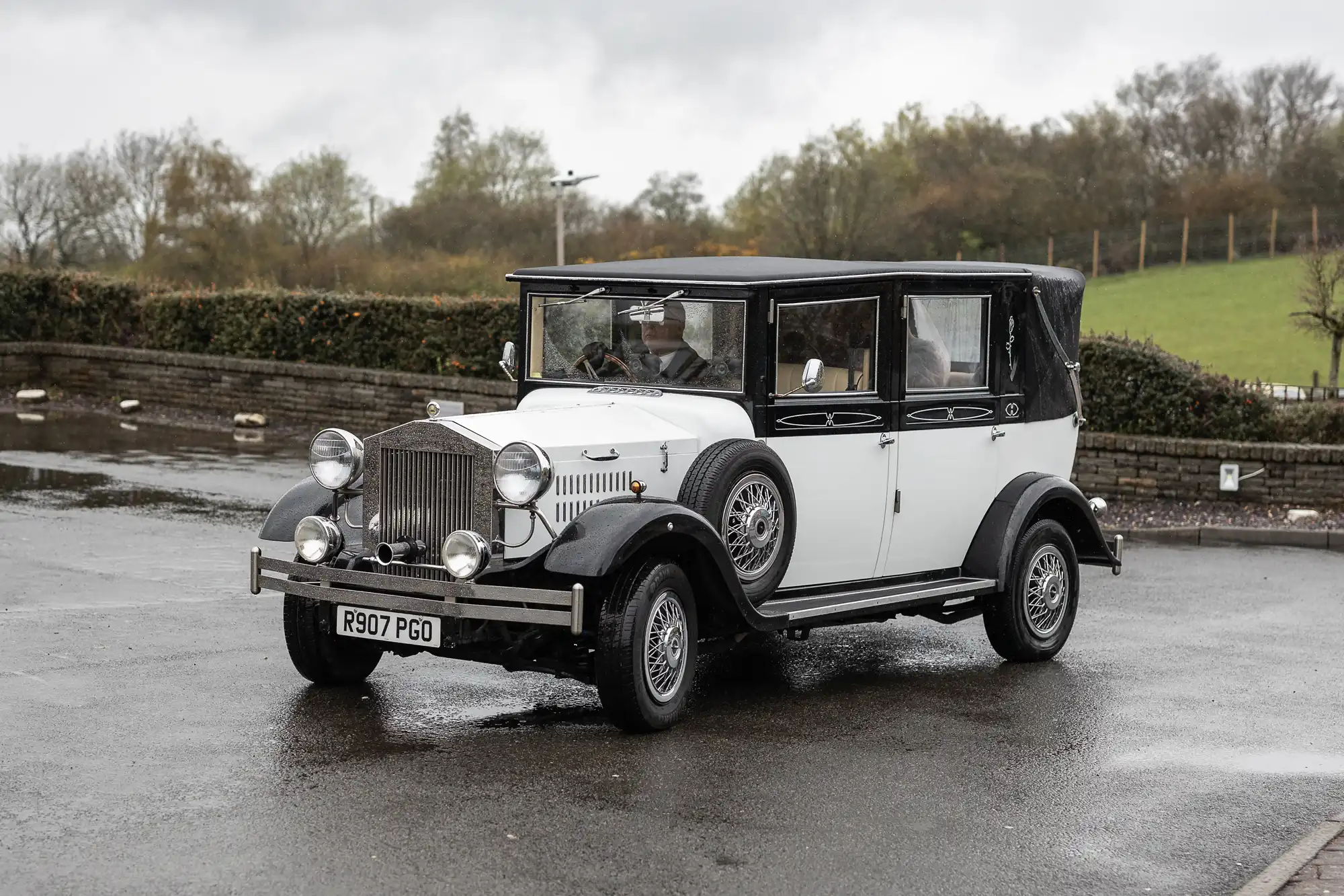 A classic white and black vintage car with a soft black roof is parked on wet pavement. The car has round headlamps, silver grille, chrome wire wheels, and registration plate R907 PGO. The background shows trees, grass, and a cloudy sky.