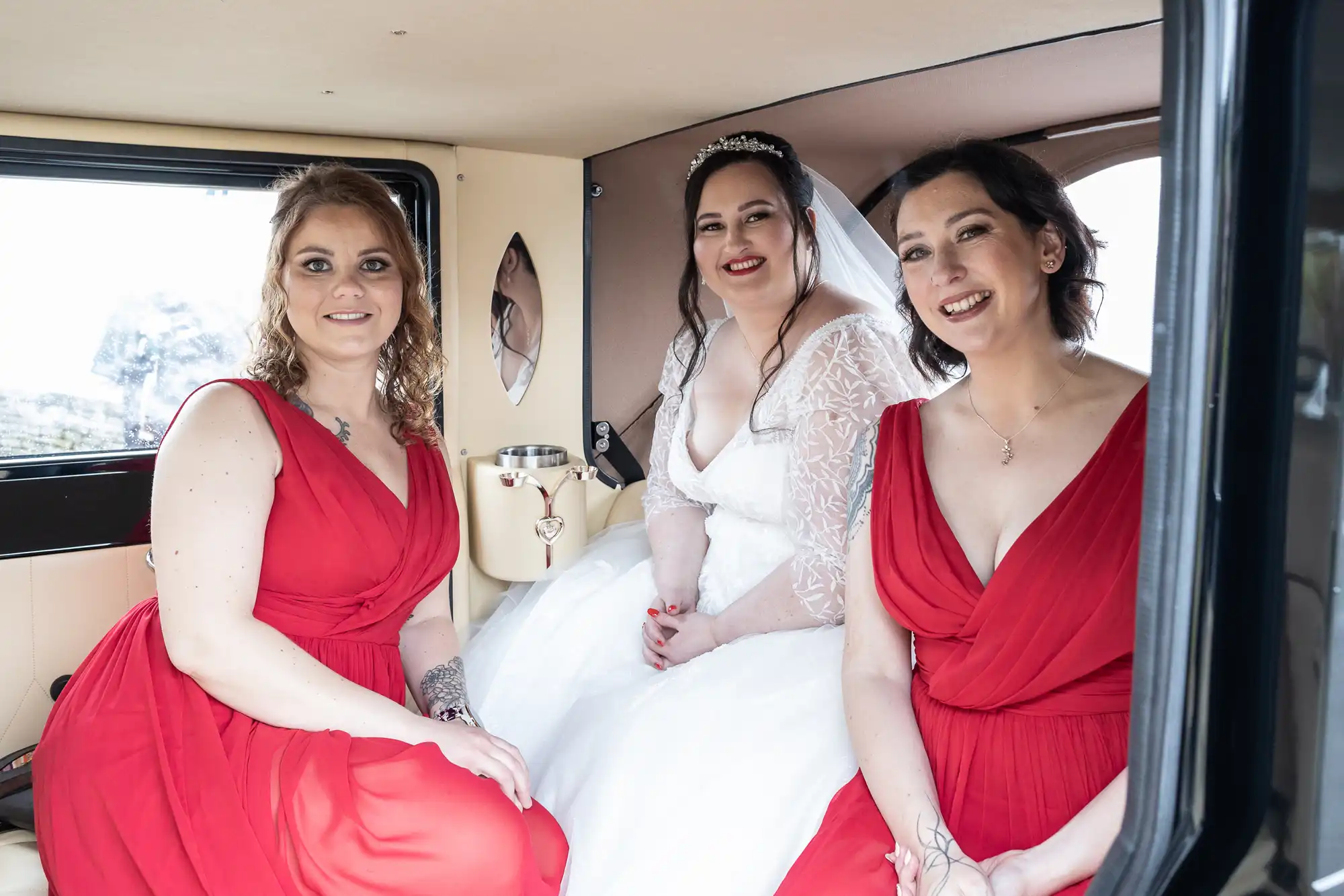 Three women sit inside a vintage car. The bride, in a white wedding dress and tiara, sits in the middle, smiling, with two bridesmaids in red dresses on either side. All three are smiling at the camera in the beige interior.
