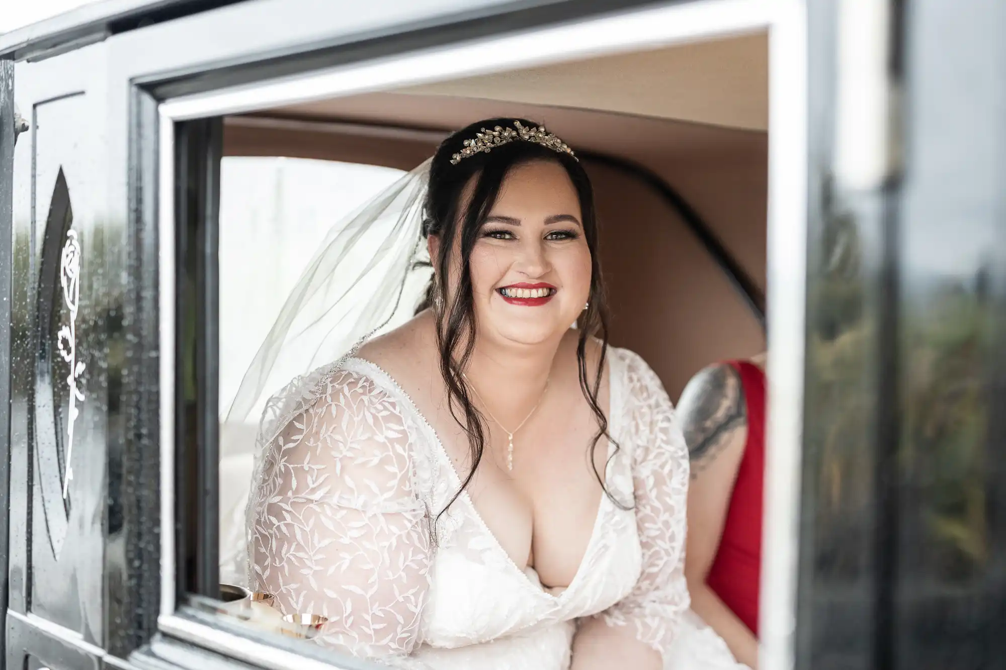 A smiling bride with dark hair, a jewelled headpiece, and a veil sits inside a vintage car. She wears a white lace wedding dress with sheer sleeves and a low neckline. Part of another person in red is visible beside her.