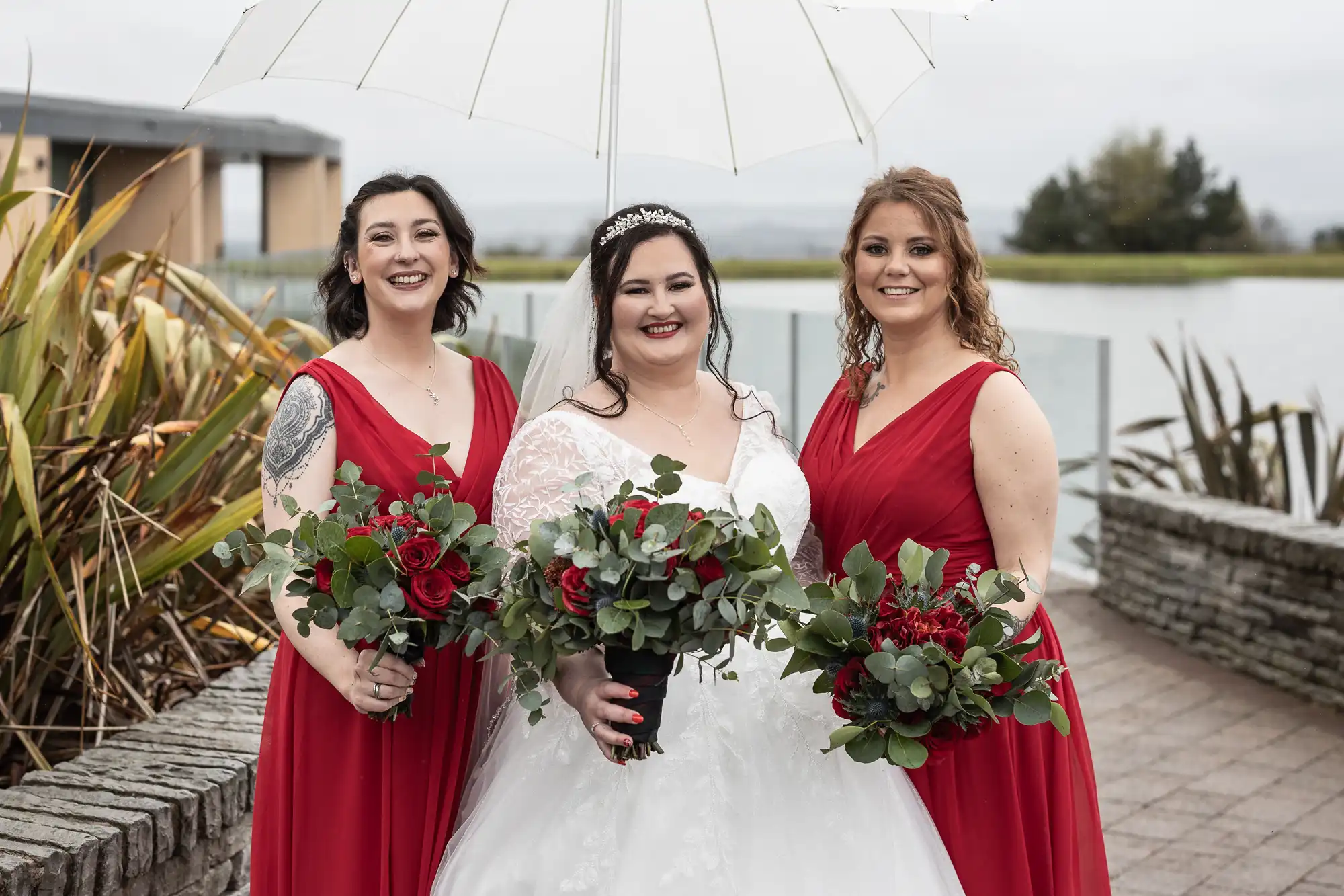 Three women stand outdoors by a lake, posing for a photo. The bride in a white dress holds a brolly and bouquet, flanked by two bridesmaids in red dresses holding matching bouquets. All are smiling, with greenery and a stone wall behind them.
