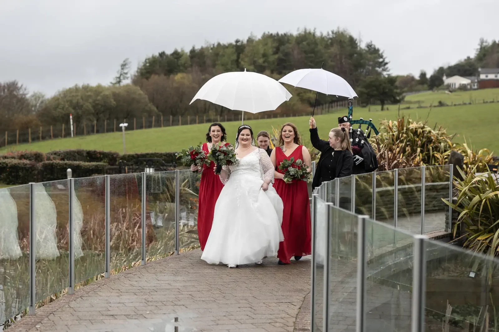 A bride in a white dress walks outdoors on a rainy day, smiling and holding a bouquet, flanked by two bridesmaids in red dresses; another person holds white umbrellas over them, with green hills and a glass fence in the background.