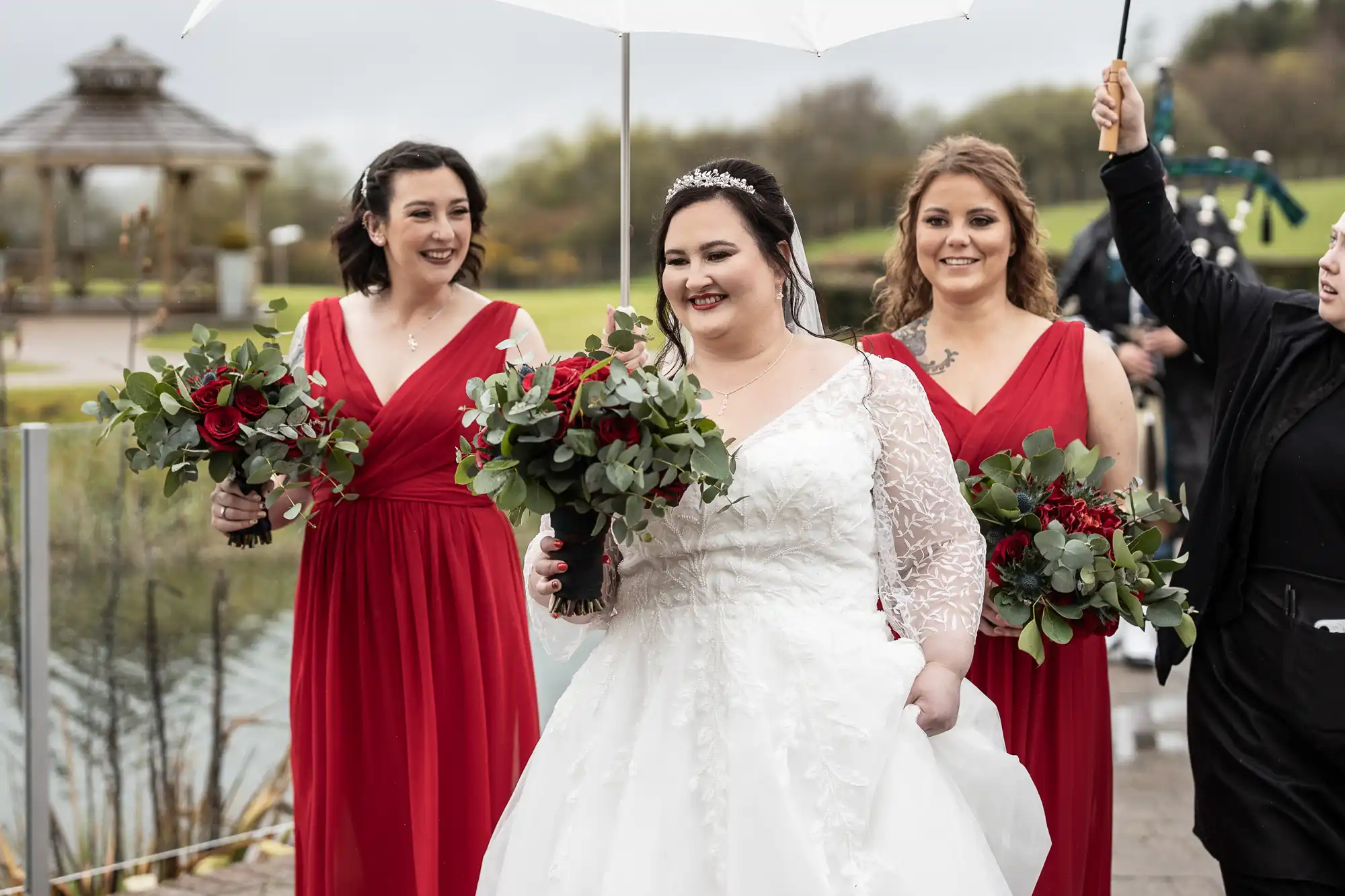 A bride in a white dress and tiara smiles while holding her gown’s skirt, flanked by two bridesmaids in red dresses holding bouquets of red roses and greenery. Another person holds a white umbrella over the bride. Trees and a gazebo appear in the background.