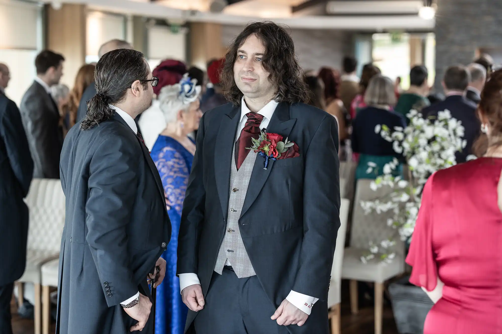 A man in a dark suit with a burgundy cravat stands indoors among a group of formally dressed people at an event. Another man in a similar suit is turned towards him. Attendees are mingling in the background, and there are elegant floral decorations.