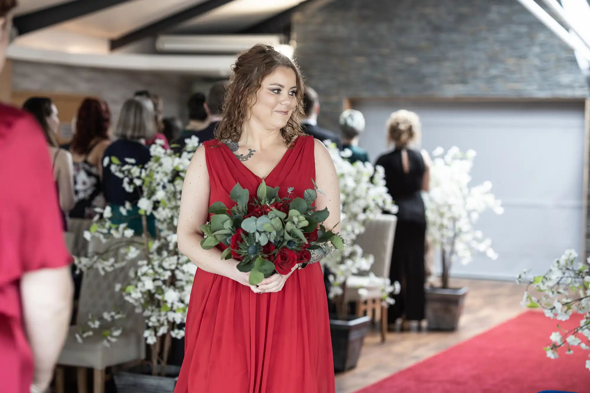 A woman in a sleeveless red dress holds a bouquet of red and green flowers, smiling as she stands indoors. Wedding guests sit and stand behind her, and white floral decorations line the aisle in a bright, modern venue.
