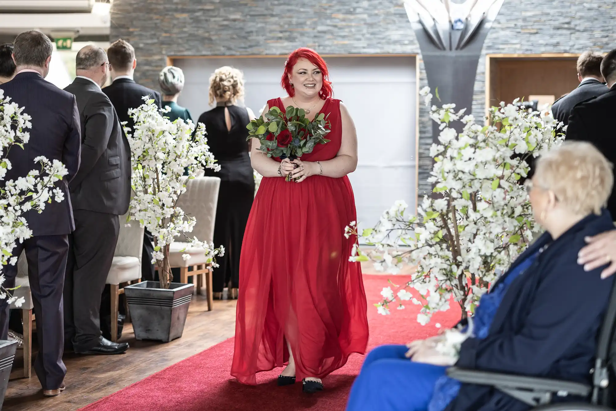A woman in a flowing red dress with bright red hair walks down a red carpet aisle, holding a bouquet of red and white flowers. Guests are seated on either side among white blossom decorations, and a person in blue is in the foreground.