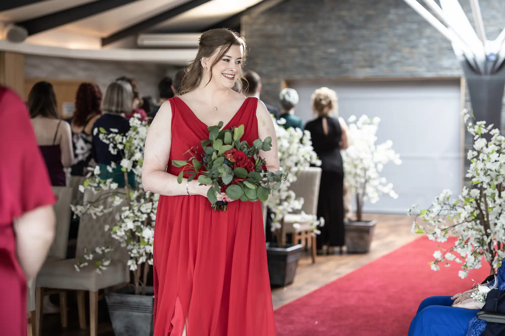 A smiling woman in a flowing red dress holds a bouquet of red and green flowers while walking down a red carpet aisle at an indoor wedding venue, surrounded by seated guests and trees with white blossoms.