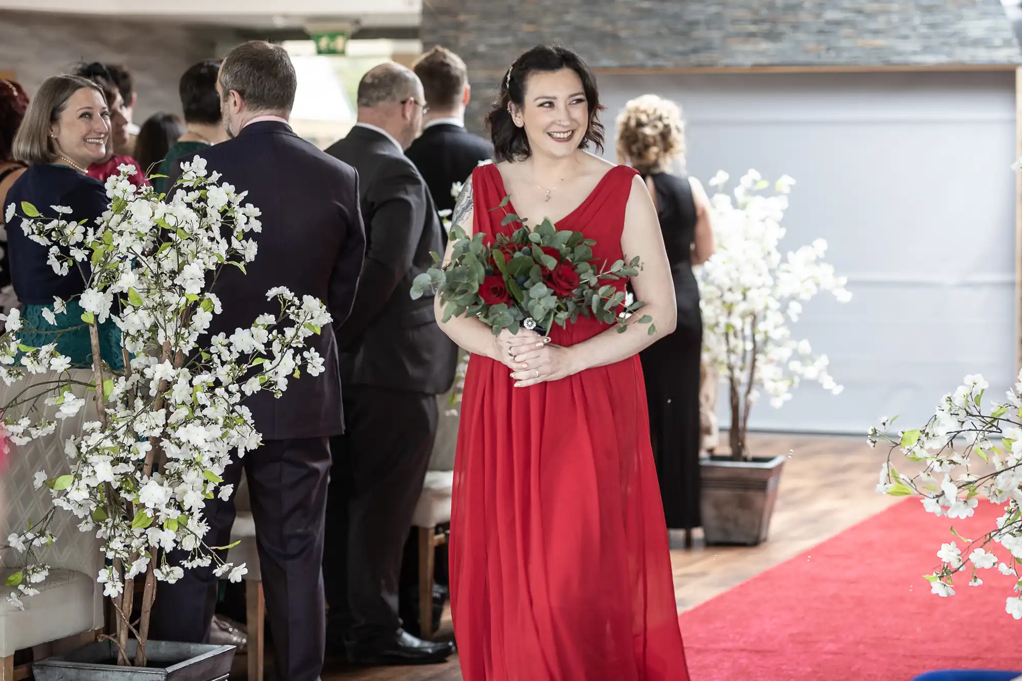 A woman in a flowing red dress holds a bouquet of red and white flowers, smiling as she walks down a red carpet aisle lined with white blossoming floral arrangements. People in formal attire watch her from both sides. The setting appears festive and elegant.