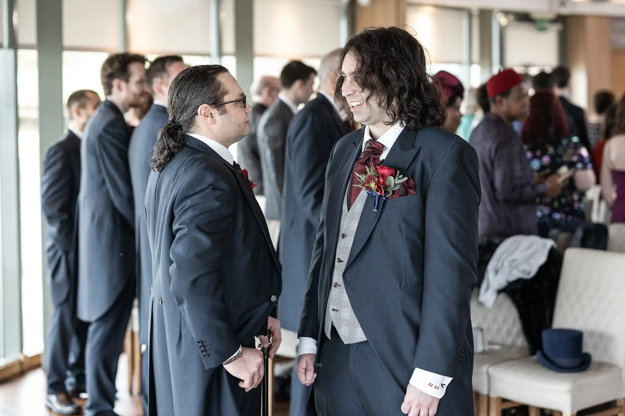 Two men in suits, one with long curly hair and the other with a ponytail and glasses, stand chatting at a formal event. Both wear buttonholes. Other guests, mostly men in suits, are mingling in the background in a bright, modern room.